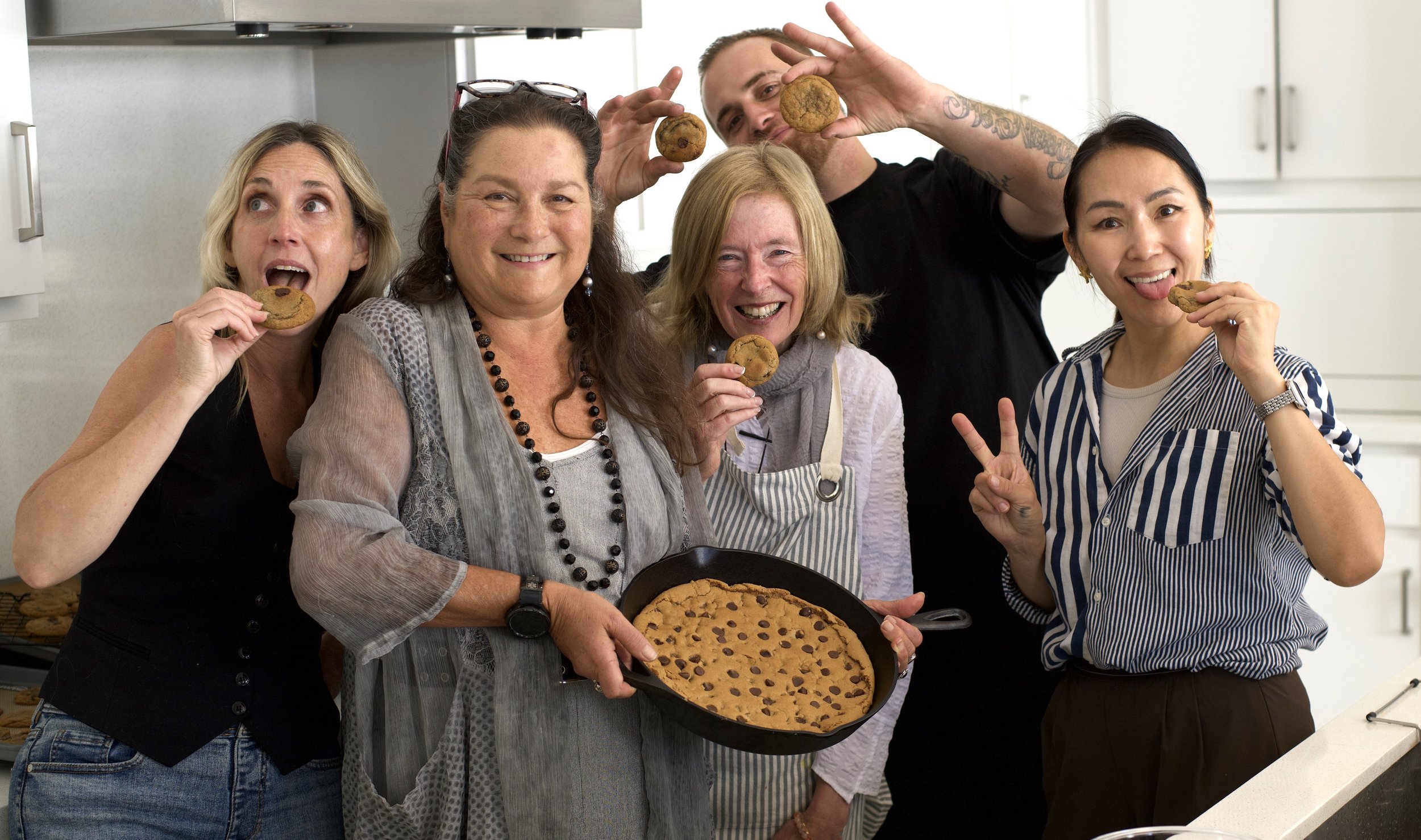 Group of five people in a kitchen, enjoying and posing with freshly baked chocolate chip cookies, with a tray and a bowl of cookies in front of them.