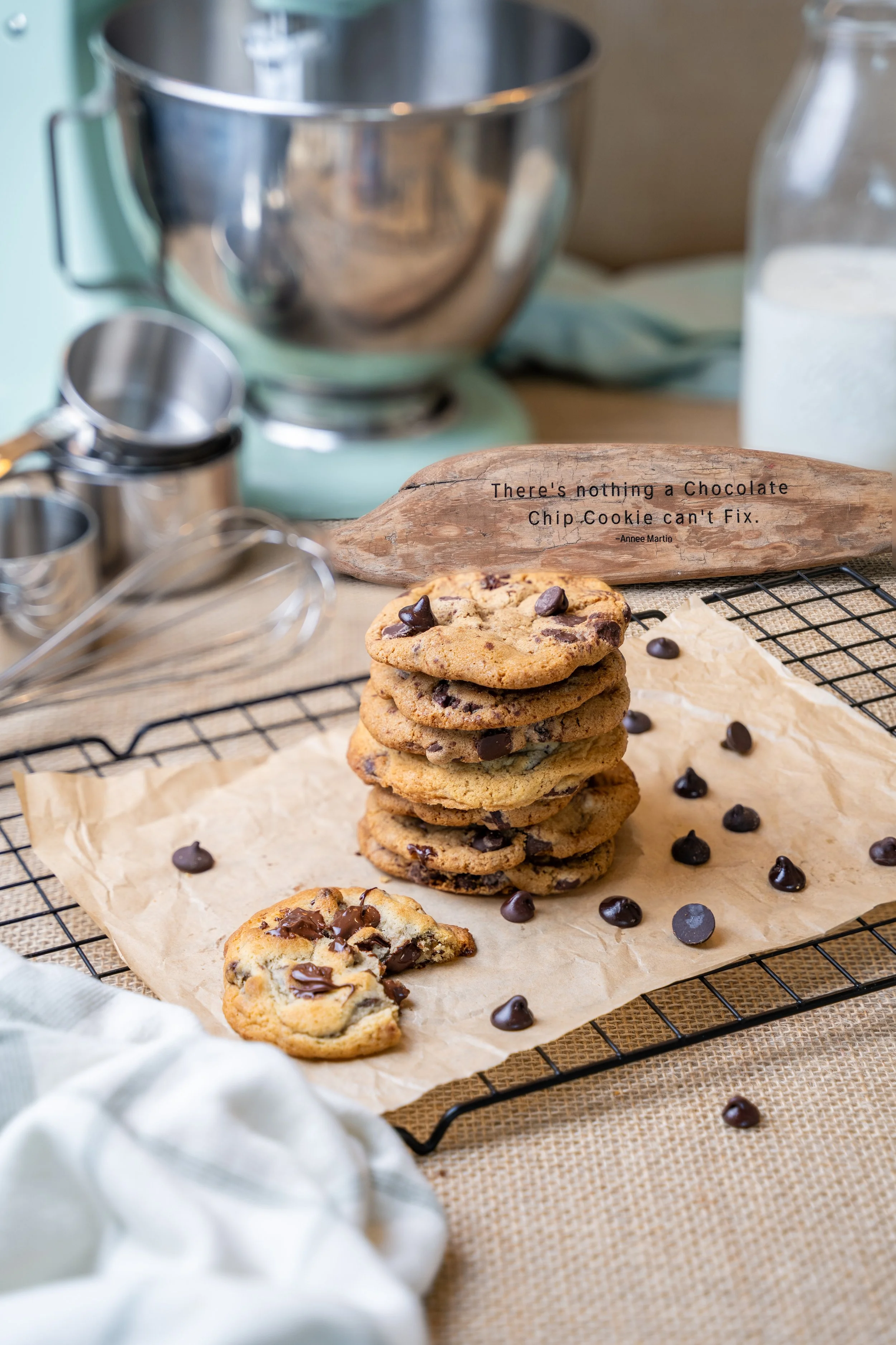 Stack of chocolate chip cookies on parchment paper, scattered with chocolate chips, on a wire cooling rack. In the background, a wooden sign reads, "There's nothing a Chocolate Chip Cookie can't Fix." with baking tools and ingredients nearby.