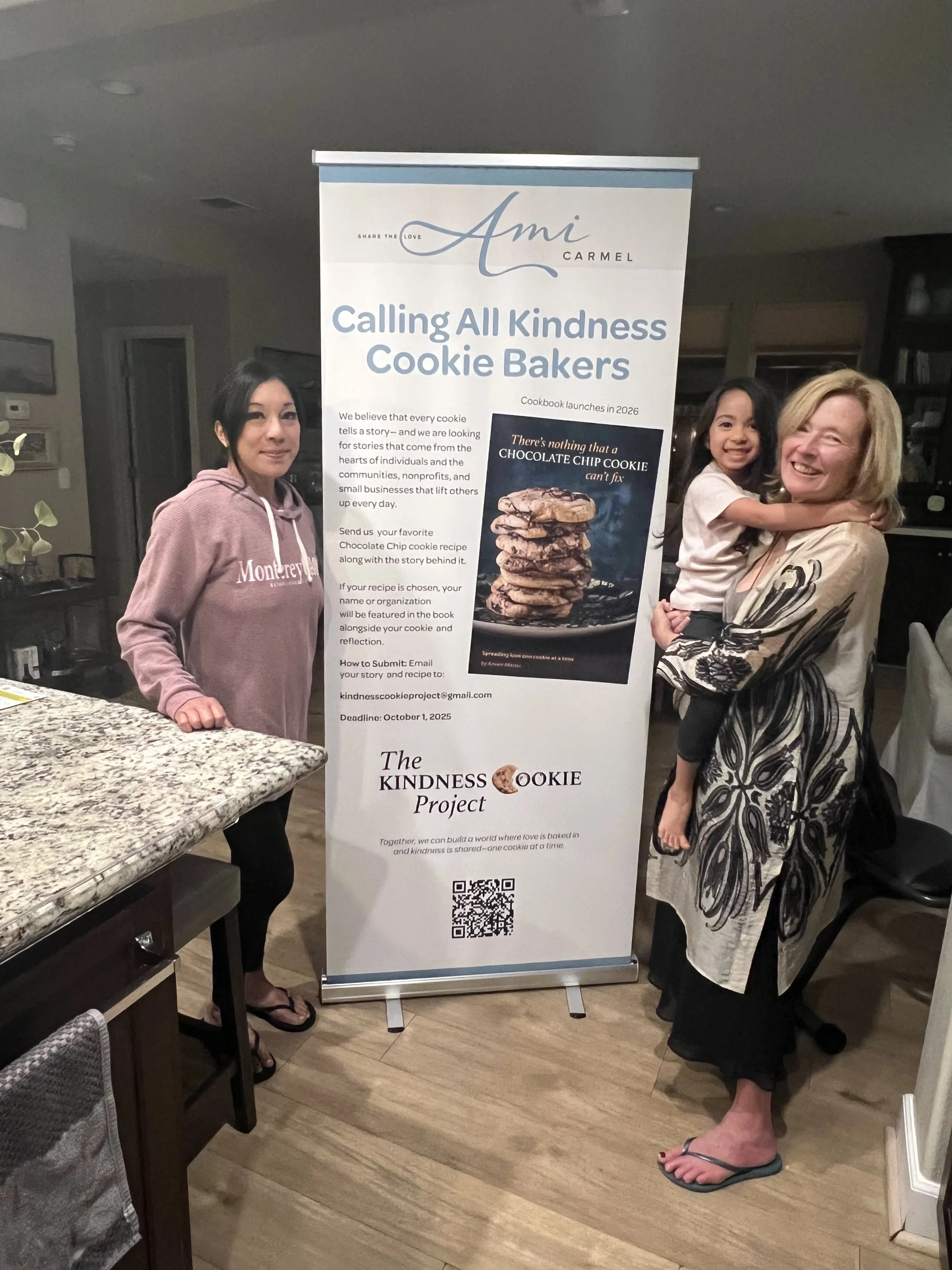 Three women standing in front of a banner for the kindness cookie project, smiling. One woman is holding a young girl. The women are dressed casually and are in a home or community center setting.