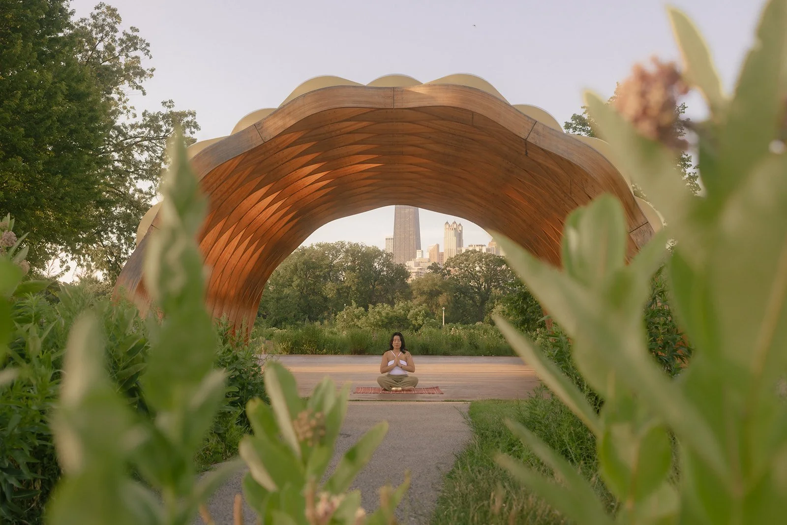 A woman practicing meditation in a park, seated on a mat under a large wooden arch structure, with city buildings visible in the background, framed by green foliage.