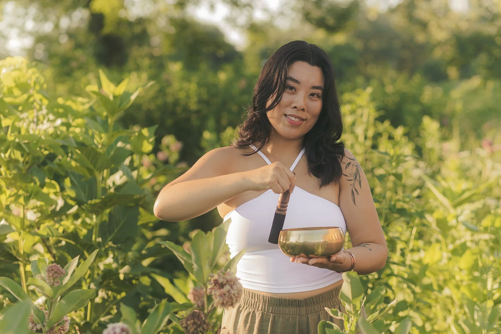 A woman holds a singing bowl and a mallet in a lush green outdoor setting, smiling at the camera.