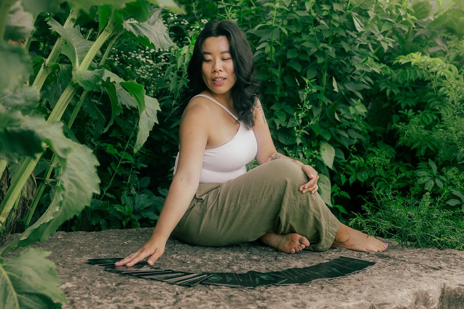 A woman with black hair and a tattoo on her arm sitting on a log in a lush green garden, with her left hand resting on a circular pattern of black leaves on the ground.