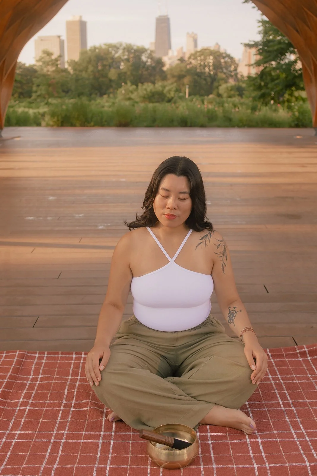 A woman sitting cross-legged on a red checkered mat outdoors, meditating with eyes closed, in front of a city skyline with trees.