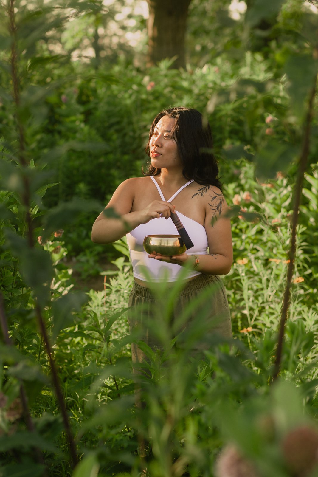 A woman with dark hair and tattoos on her shoulder, wearing a white top and brown pants, stands amidst green foliage in a garden, holding a singing bowl and a wooden mallet, with sunlight shining on her face.