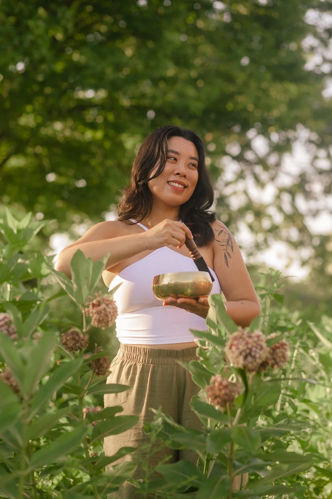 A woman smiling outdoors among green plants, holding a singing bowl with a mallet.