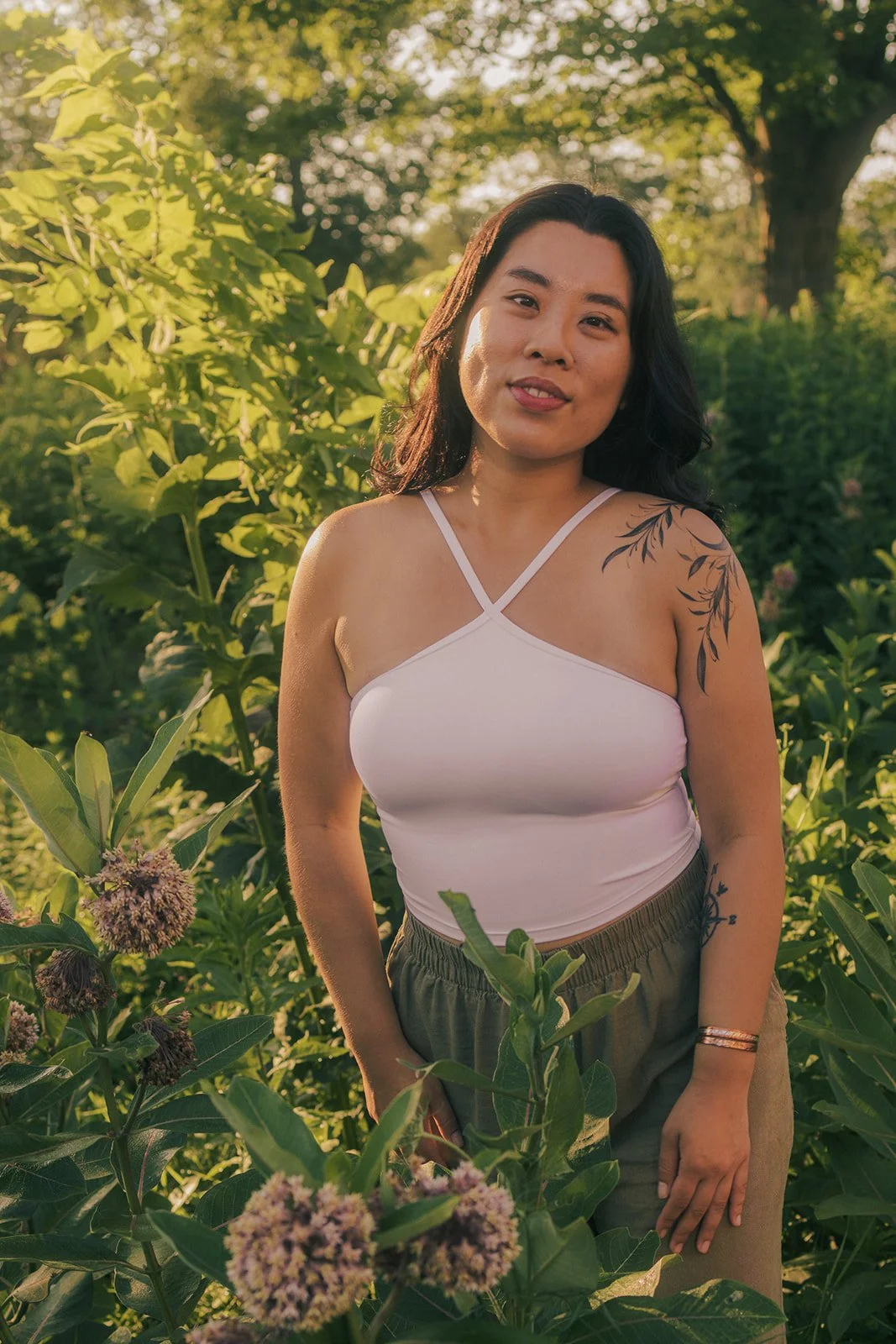 A woman with dark hair and tattoos on her shoulder and arm, wearing a white spaghetti strap top and gray pants, standing among green foliage with sunlight filtering through the trees in the background.