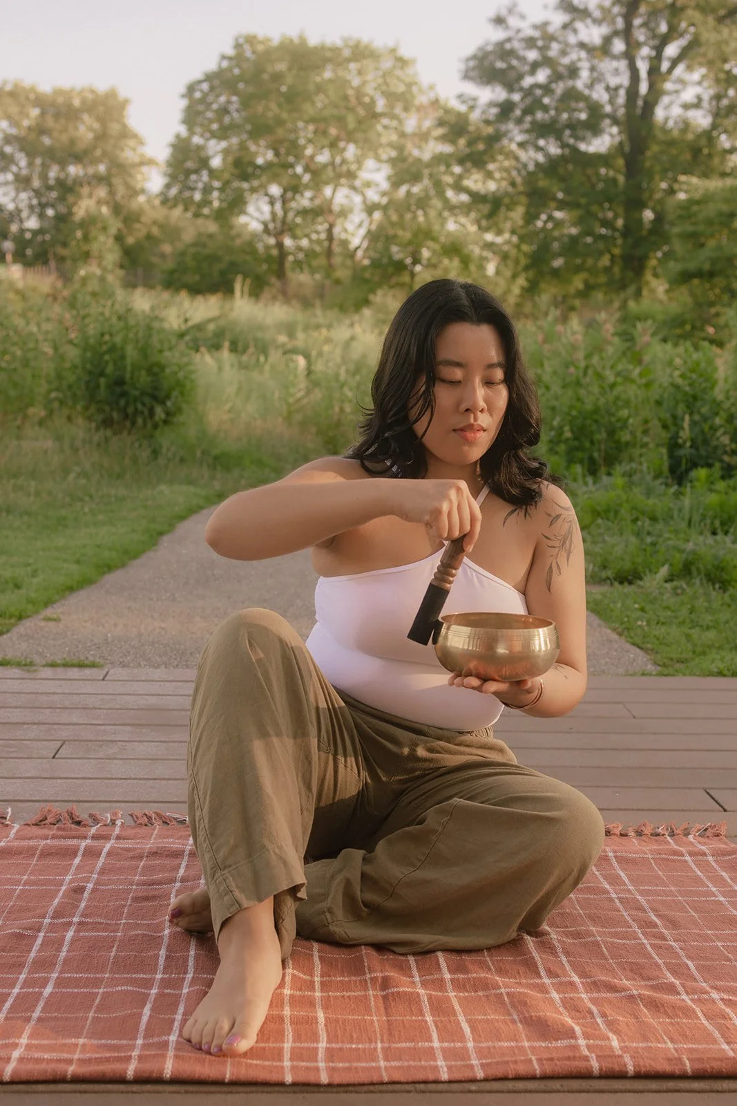 A woman sitting cross-legged on a blanket outdoors, holding a singing bowl and a mallet, with trees and a path in the background.