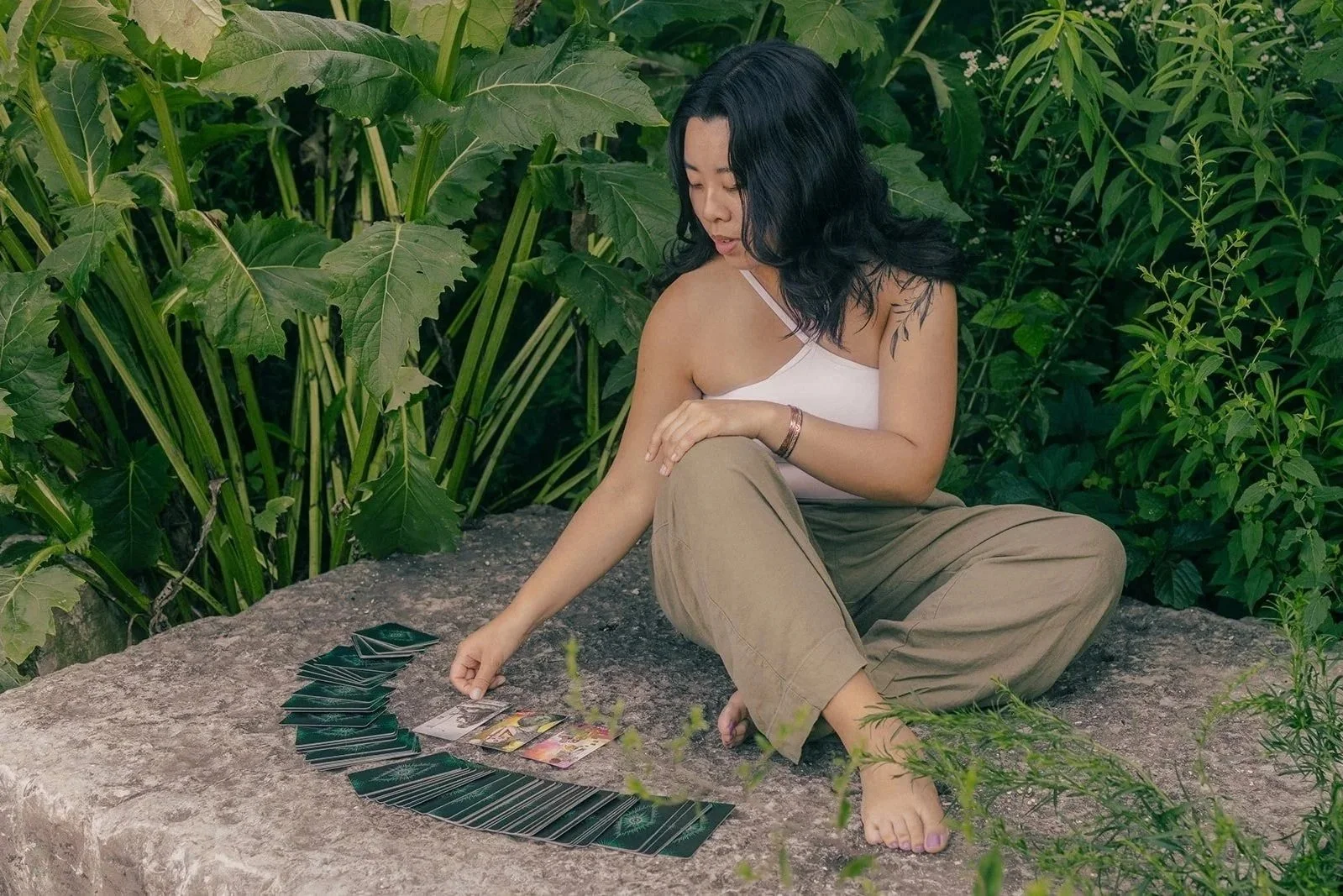 A woman with black hair wearing a white tank top and khaki pants sitting on a large rock surrounded by green plants, playing a card game with cards spread out on the rock.