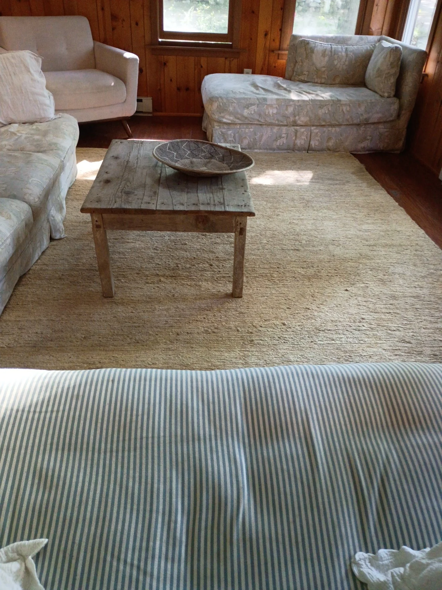 View of a cozy living room with wooden walls, two sofas — one white and one floral patterned — a rustic wooden coffee table with a decorative bowl, a rug, and windows letting in natural light.