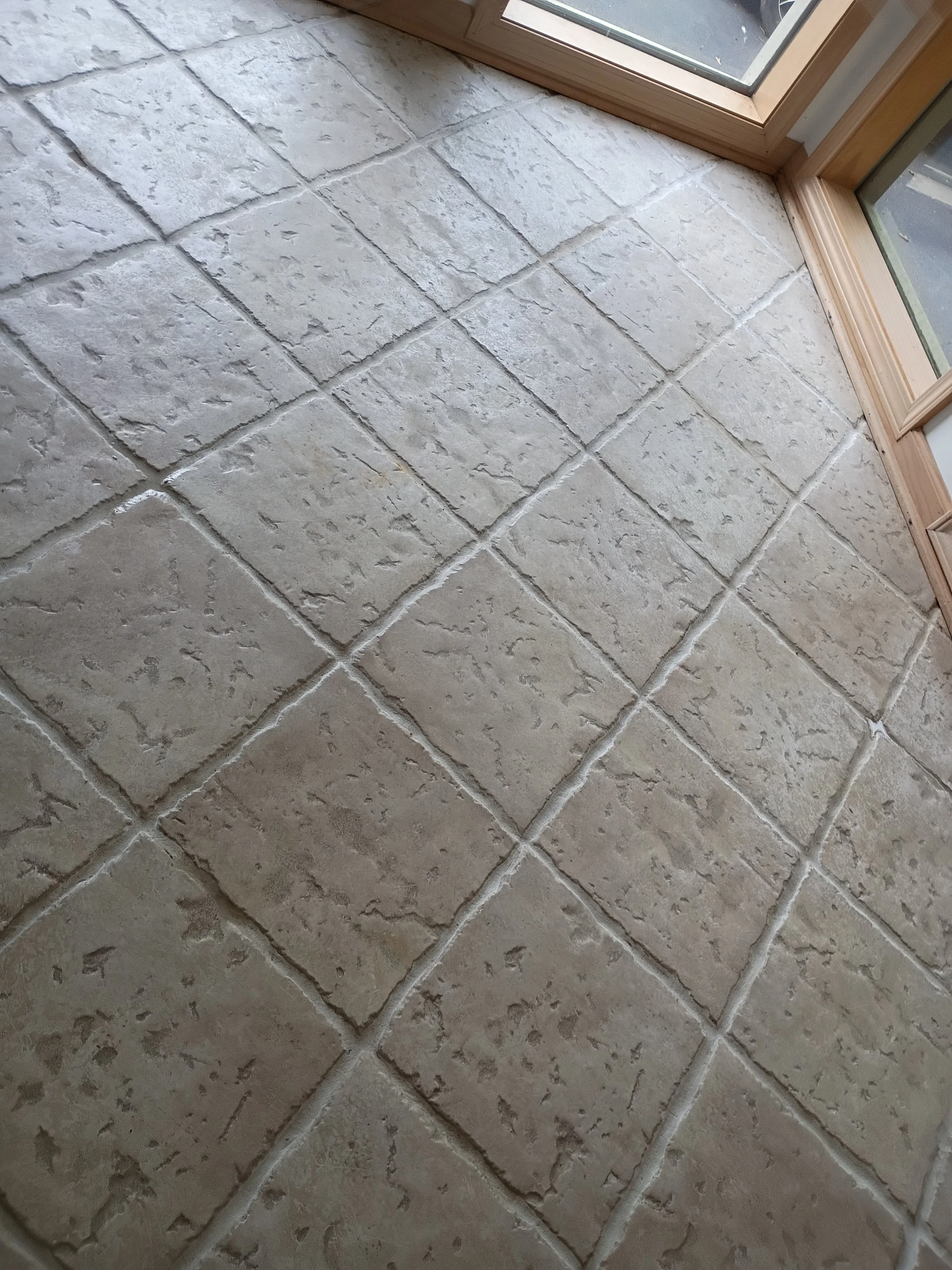 Close-up view of a tiled floor with rectangular beige tiles, where the tile edges are outlined with white grout, next to a wooden door and window frame.