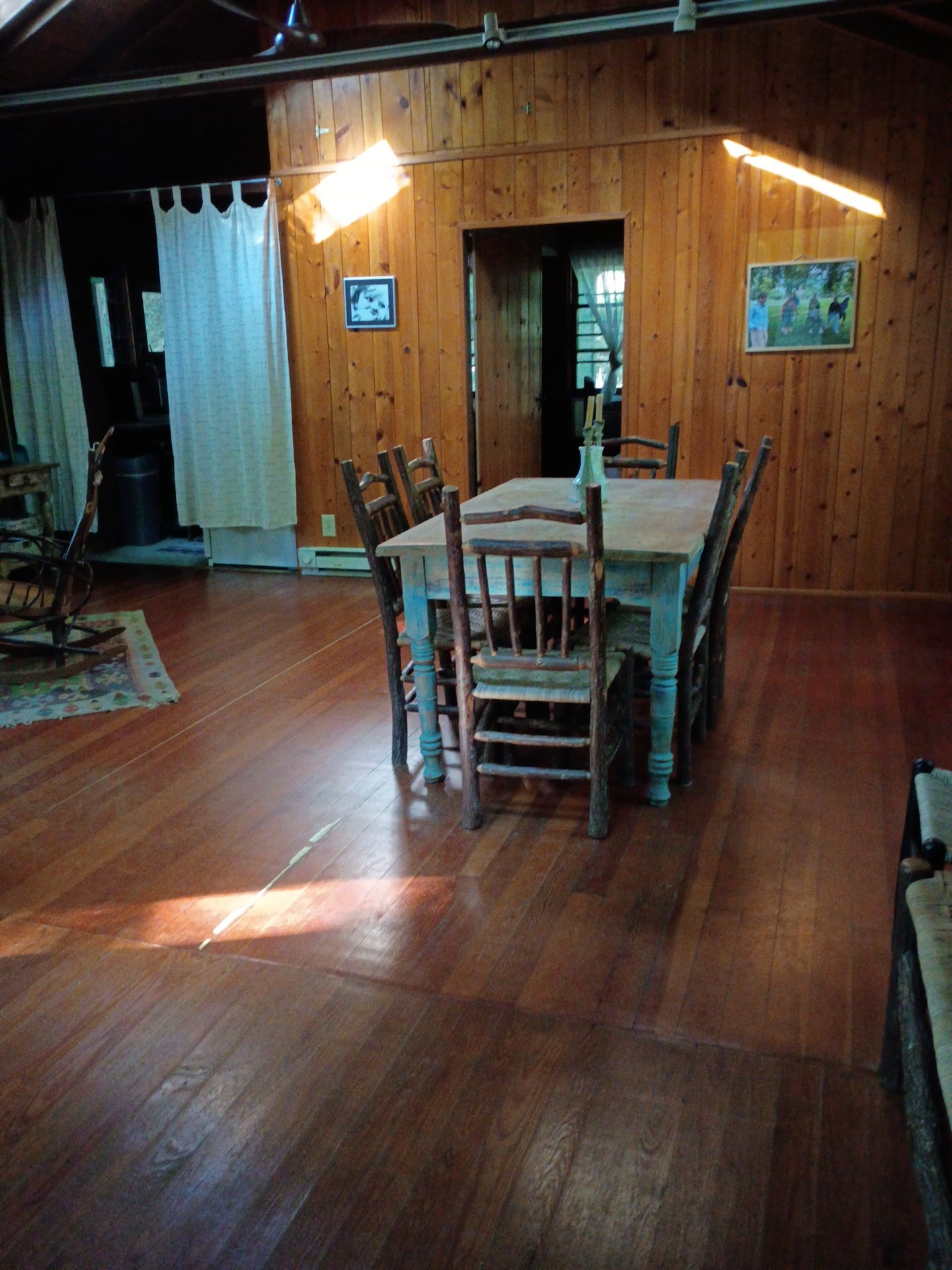 A dining room with wood-paneled walls and hardwood floors. A rustic wooden table with chairs is in the center, with a clear glass vase containing flowers on top. Sunlight streams in from the right side, creating bright streaks and shadows on the wall.