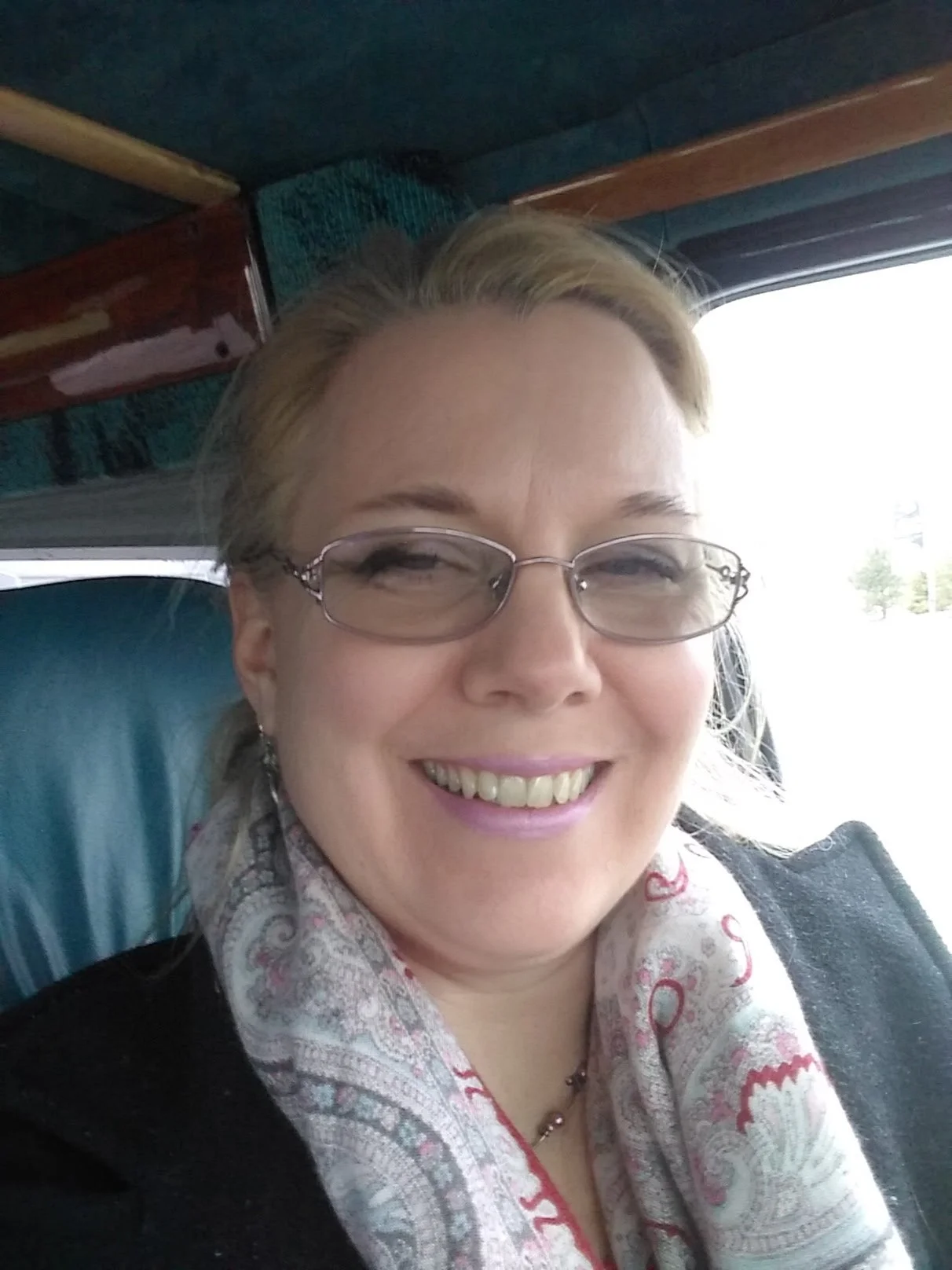 A woman with blonde hair, glasses, and light lipstick smiling, sitting in a vehicle with a blue seat and window showing outside scenery.
