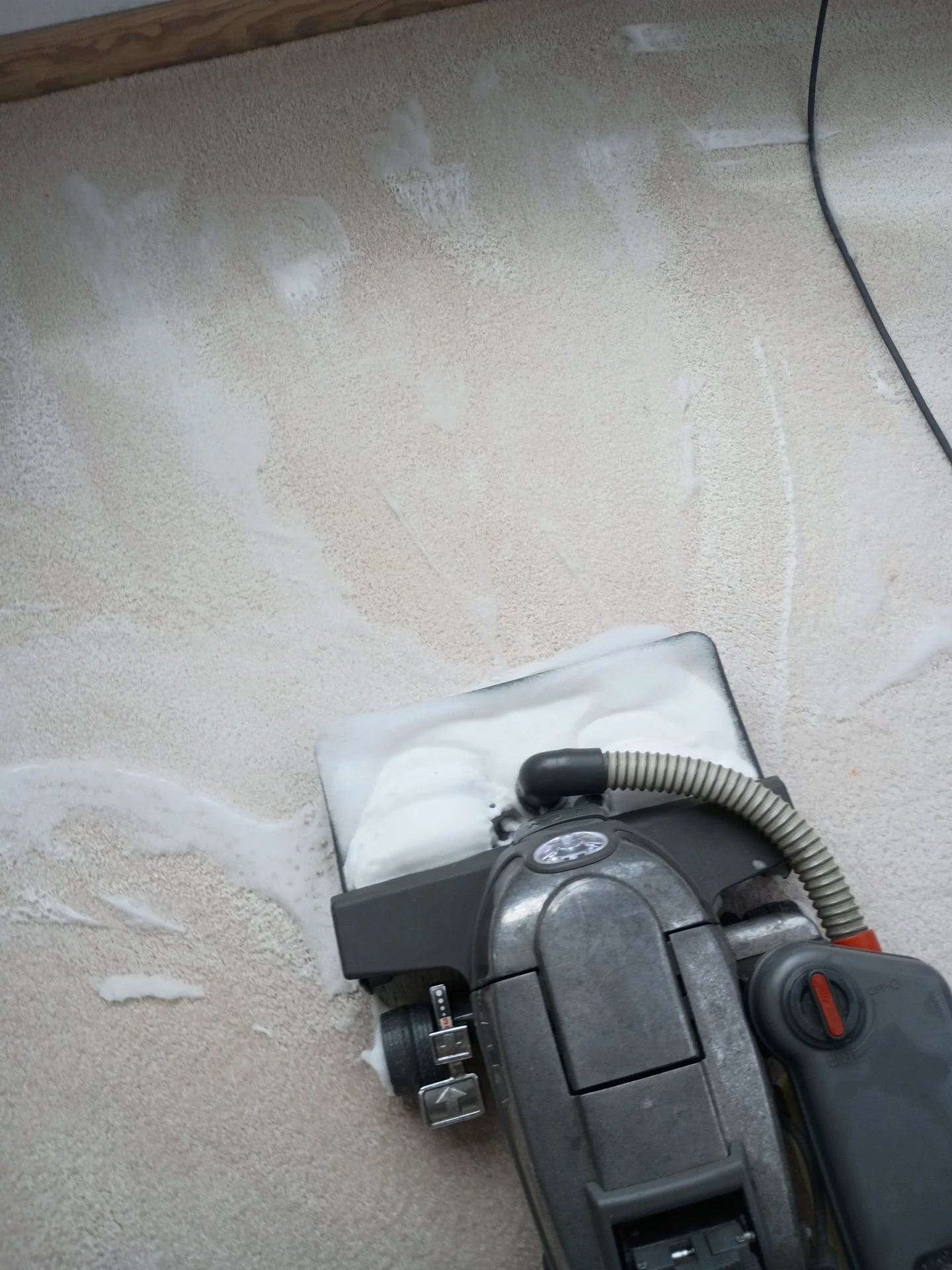 A handheld vacuum cleaner on a beige carpet, with dust and debris around it, and a power cord extending to the right.