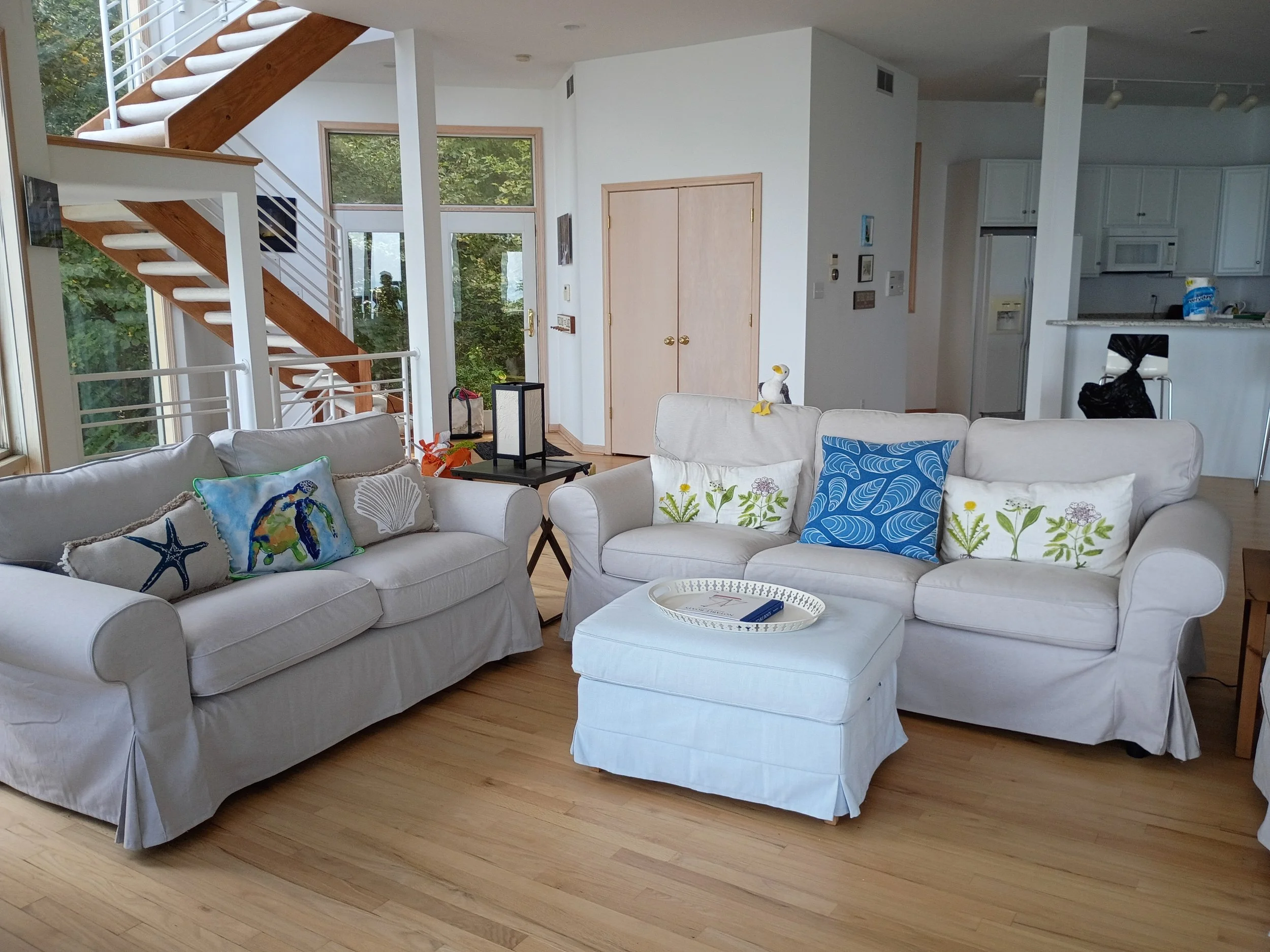 Living room with white sofas, decorative pillows, a square ottoman, a staircase with wooden steps and white railings, a large window, and a view of green trees outside.