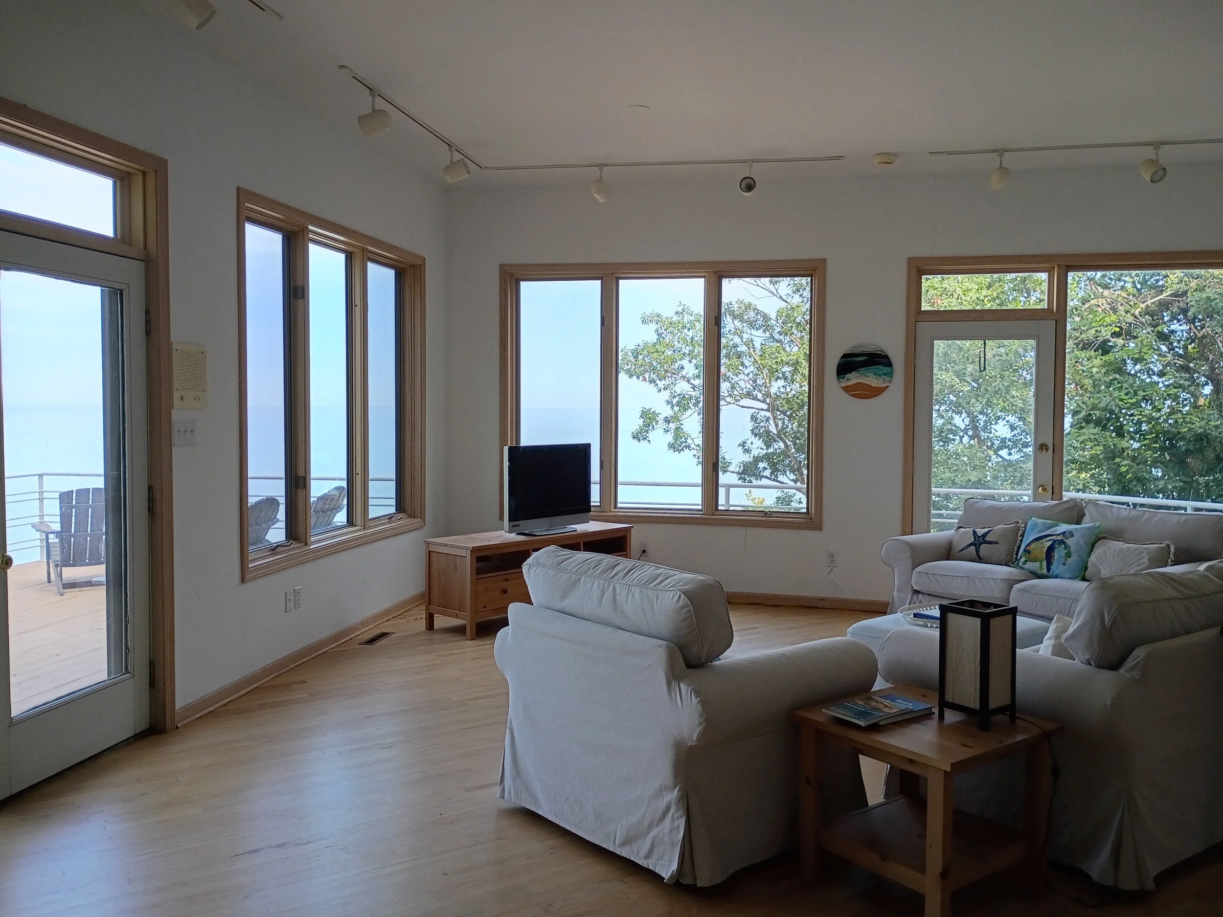 Living room with large windows showing trees and an ocean view, white sofa and chairs, a wooden coffee table with magazines, a TV on a wooden stand, and framed artwork on the wall.