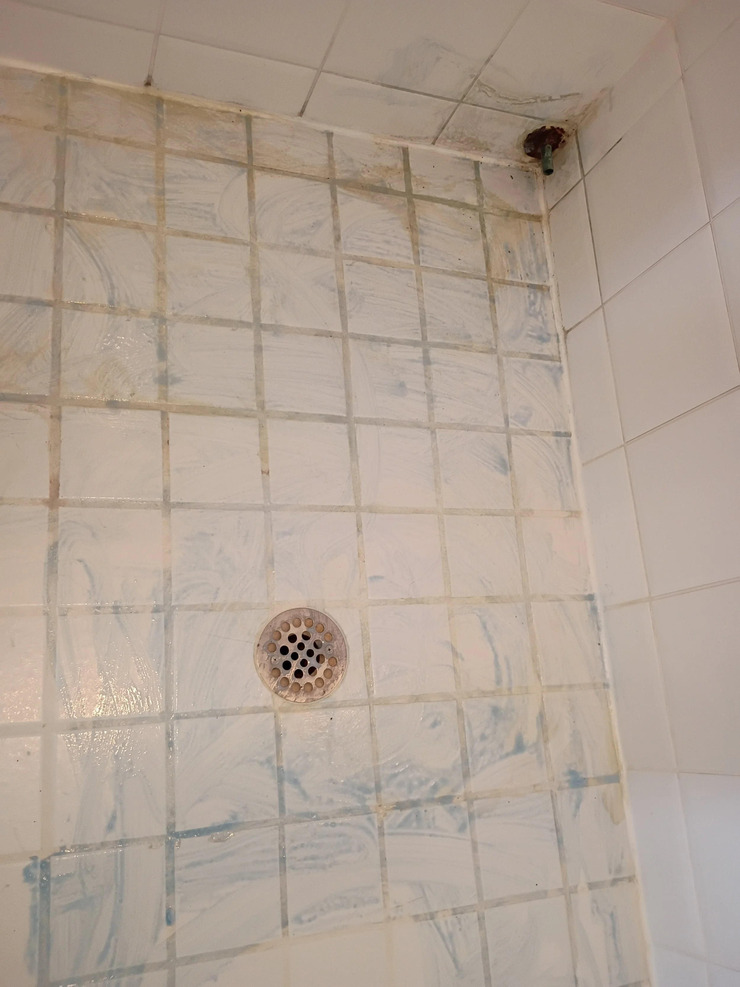 The image shows a corner of a tiled bathroom or shower with a drain and a rusted pipe fitting in the corner.