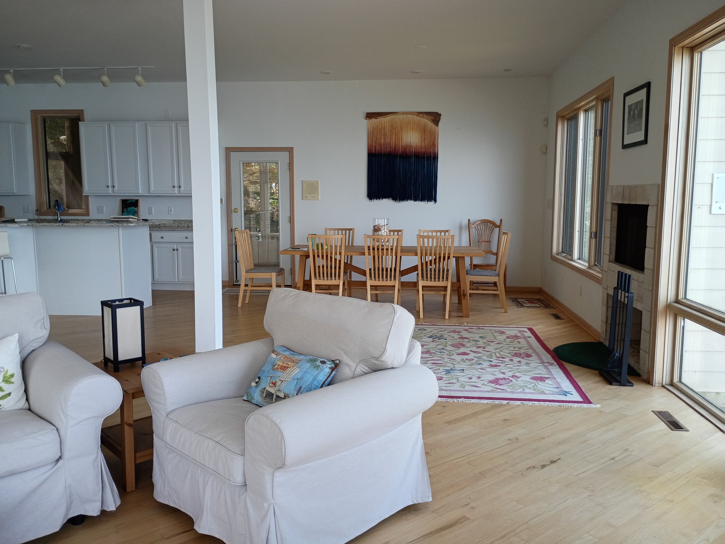 A cozy living room with white sofas, a wooden dining table with chairs, a colorful woven wall hanging, large windows with wooden frames, and a fireplace.