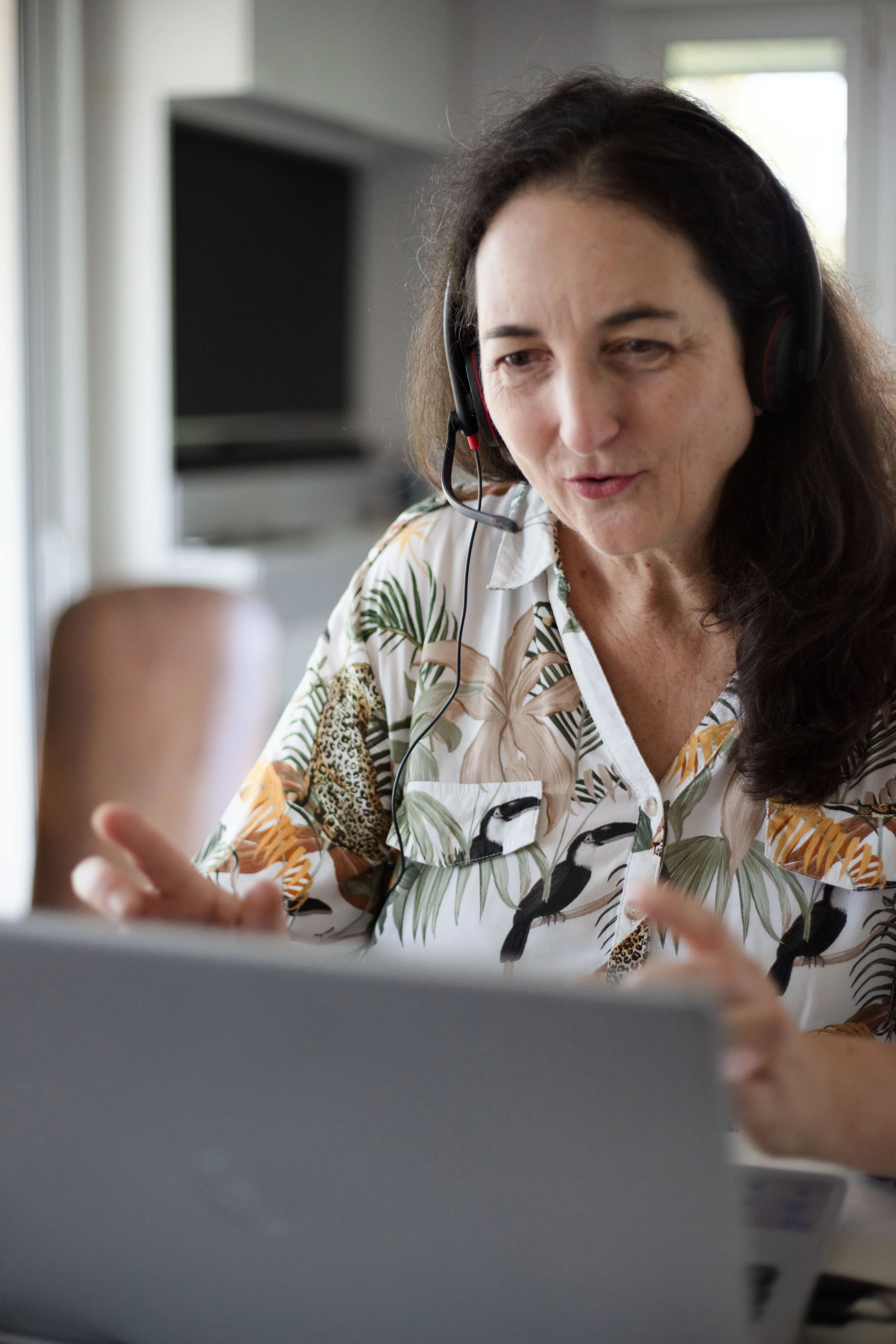 Une femme avec des cheveux longs, portant un casque avec microphone, regarde un écran d'ordinateur. Elle porte une chemise avec un motif de feuillages et d'animaux. Elle semble concentrée, peut-être en communication ou en travail à distance.
