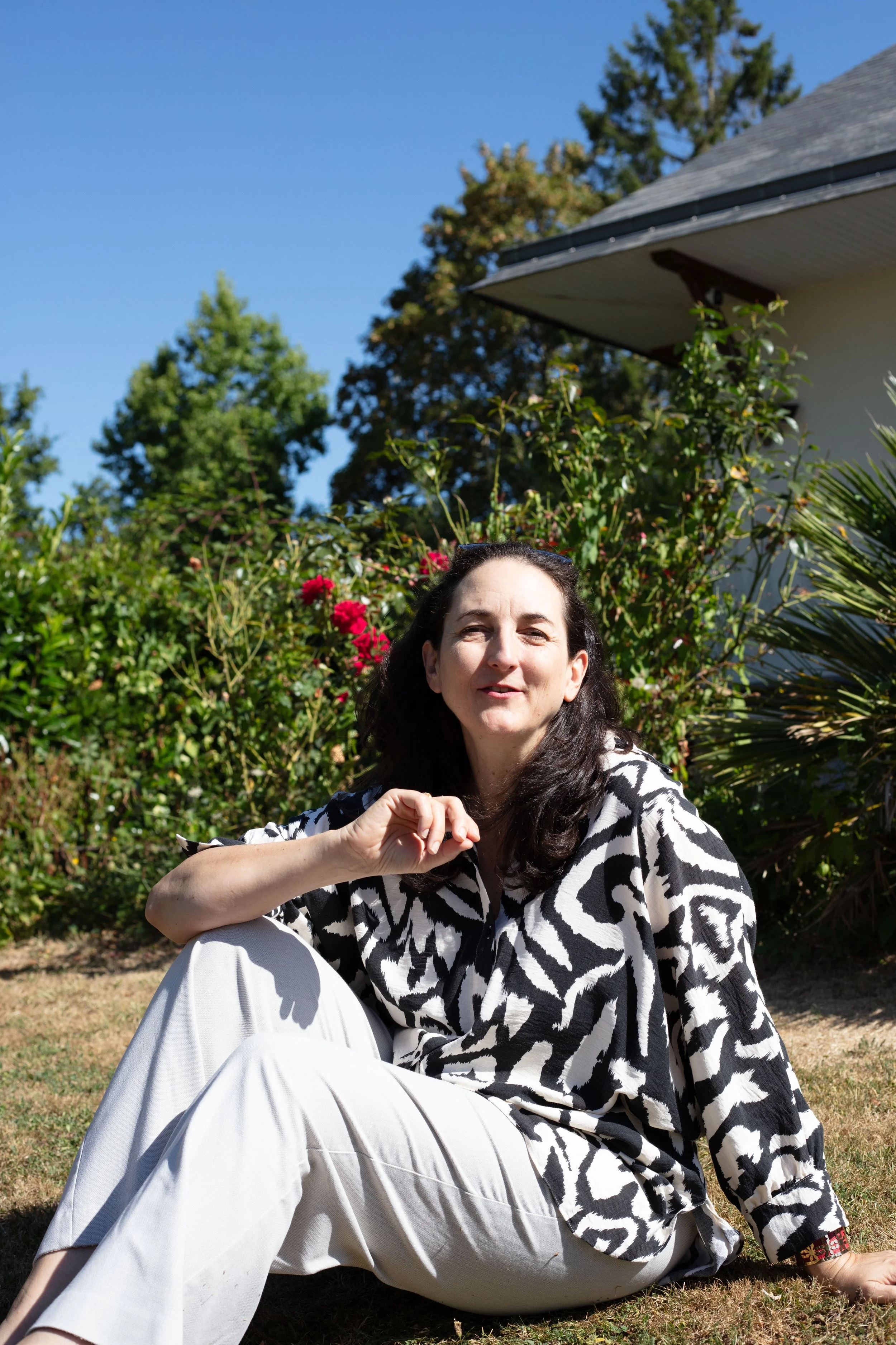 Une femme assise dans un jardin ensoleillé, portant un vêtement à motif zébré noir et blanc, avec des arbres en arrière-plan et un ciel bleu clair.