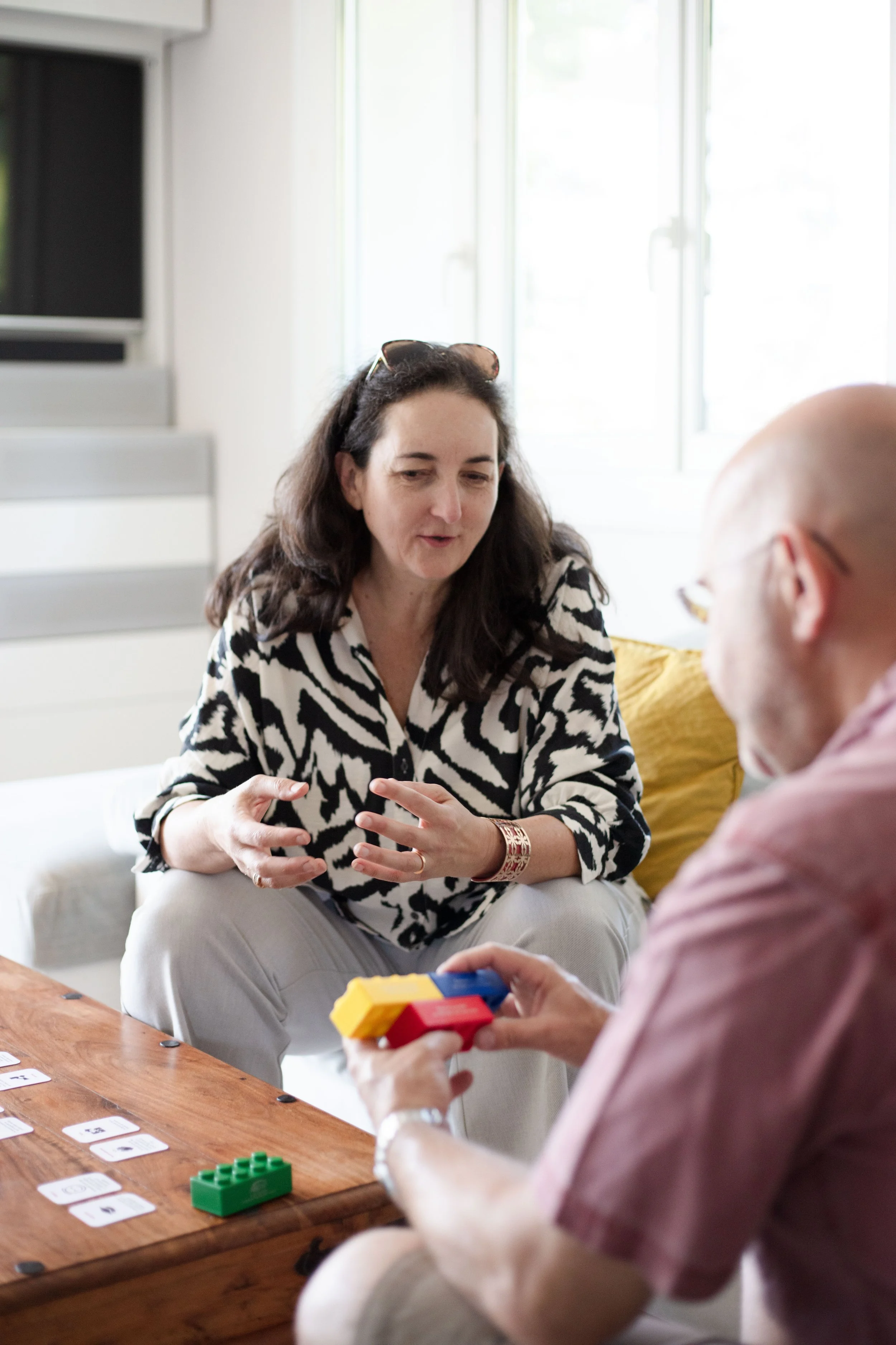 Deux personnes jouent aux jeux de société dans une pièce ensoleillée. La femme est assise, portant un chemisier à motifs animaux et parlant avec l'homme, qui tient des blocs de construction LEGO. La table a des cartes à jouer et des pièces de jeu.