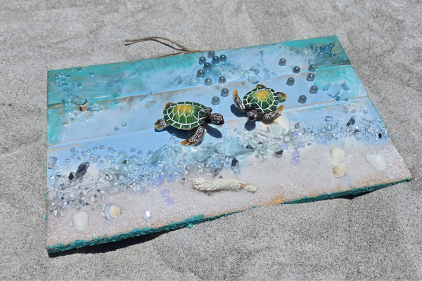 Decorative beach scene with two small turtle figurines on a wooden board painted with ocean waves, surrounded by seashells, beads, and glitter.