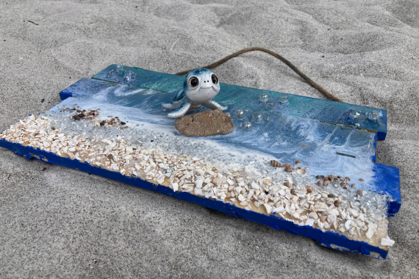 A toy seagull on a makeshift beach made of a plastic board, shells, and rocks on sandy ground.