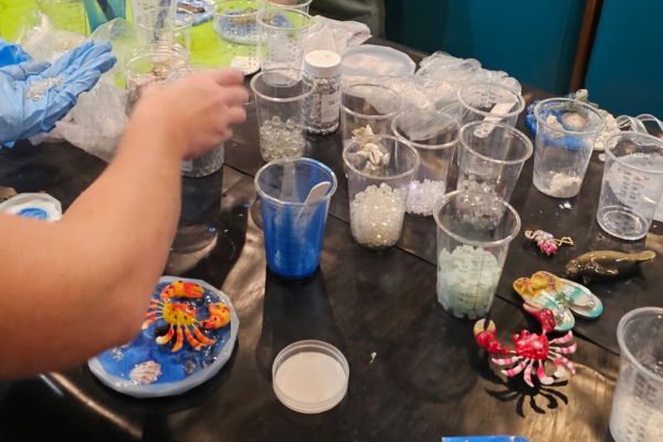A table with various containers filled with beads and small craft supplies, and some decorated crab and starfish figurines. A person�s arm is reaching towards the supplies.