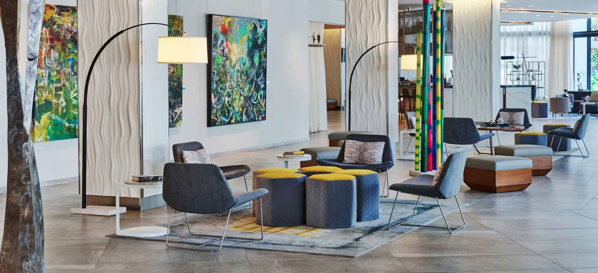 Modern hotel lobby with abstract artwork on white textured walls, multiple chairs and ottomans arranged around small tables, colorful vertical sculptures, large windows with curtains, and contemporary decor.