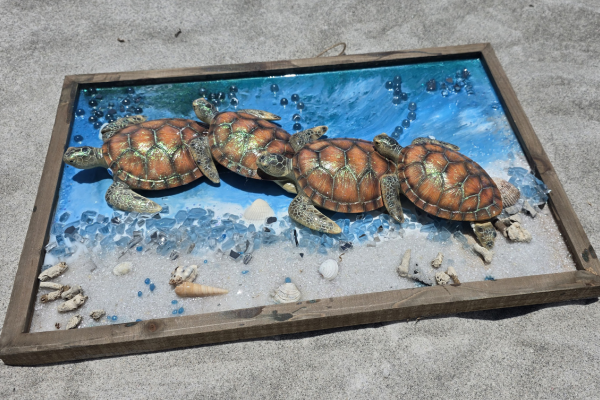 Four small turtles on a sandy beach within a wooden frame that resembles a miniature pool, with water, blue beads, shells, and small rocks.