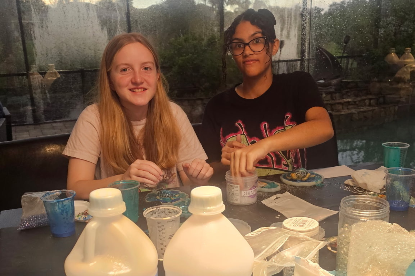 Two young girls sit at a table with arts and crafts supplies, creating ceramic turtle decorations, in a room with large windows showing an outdoor garden with rain.
