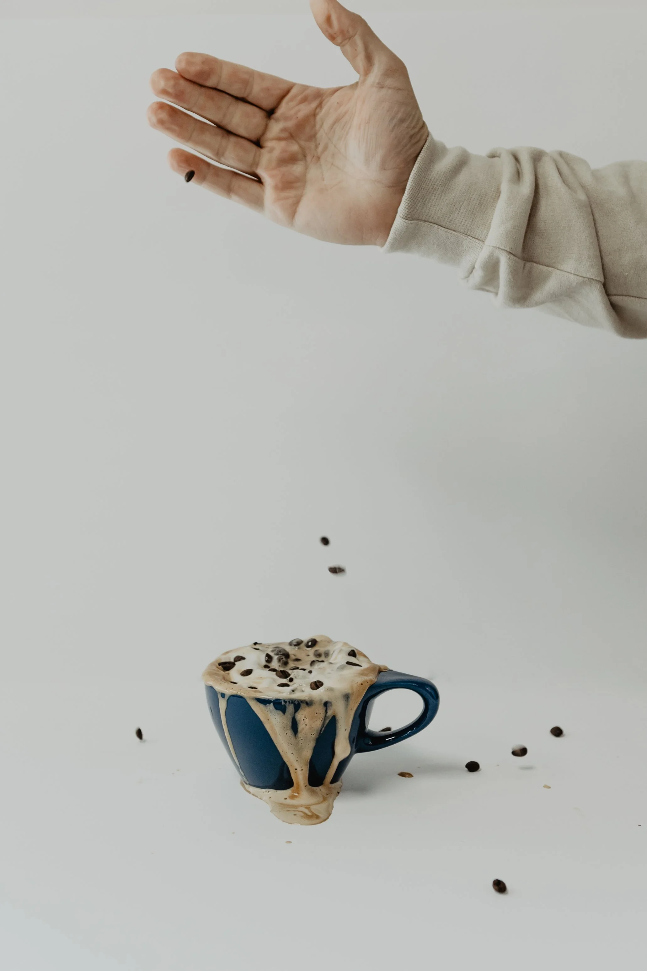 A hand is sprinkling coffee beans over a blue mug filled with a creamy beverage topped with chocolate chips, against a plain white background.