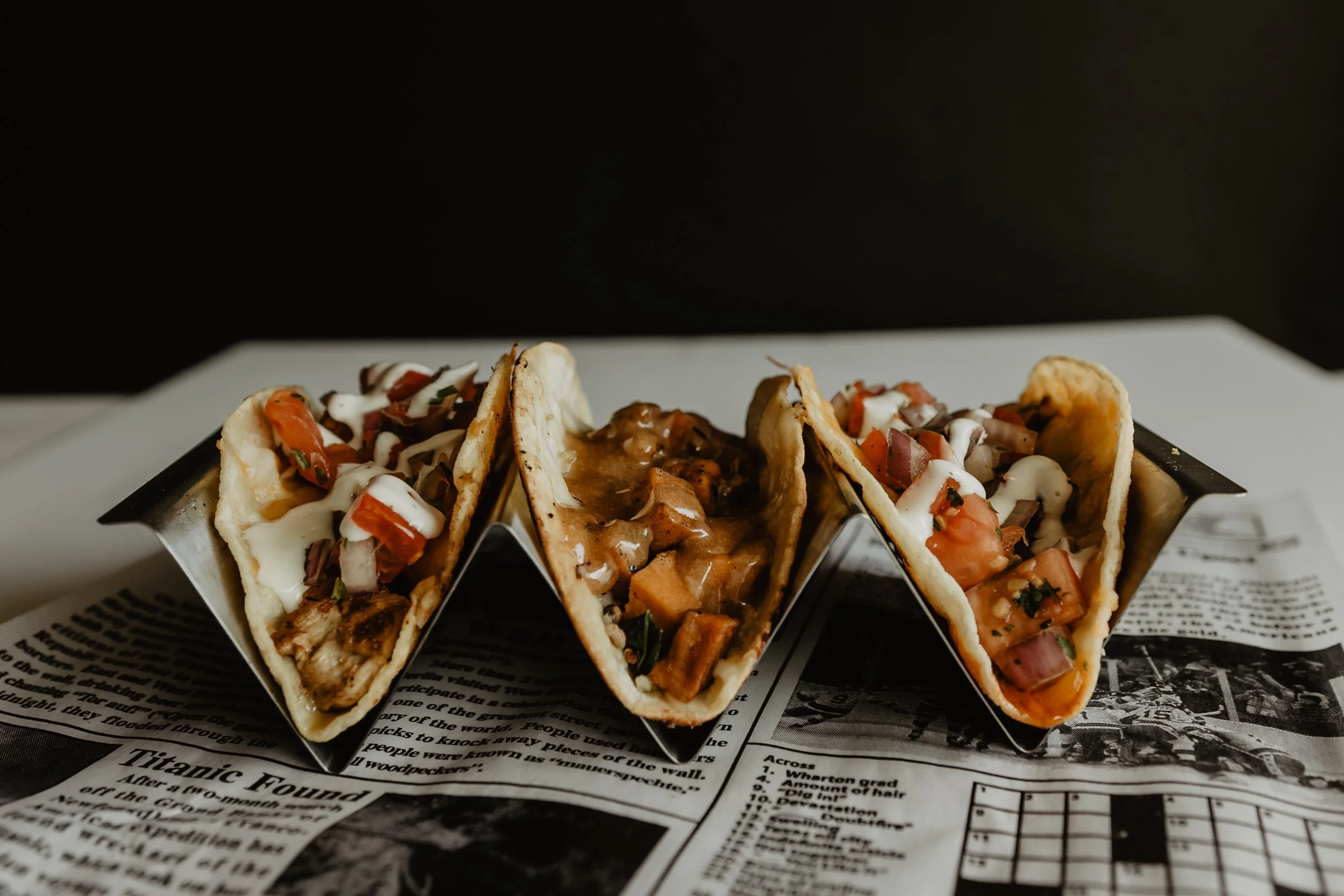Three tacos filled with chopped tomatoes, onions, and cilantro, with a drizzle of white sauce, on a tray lined with newspaper.