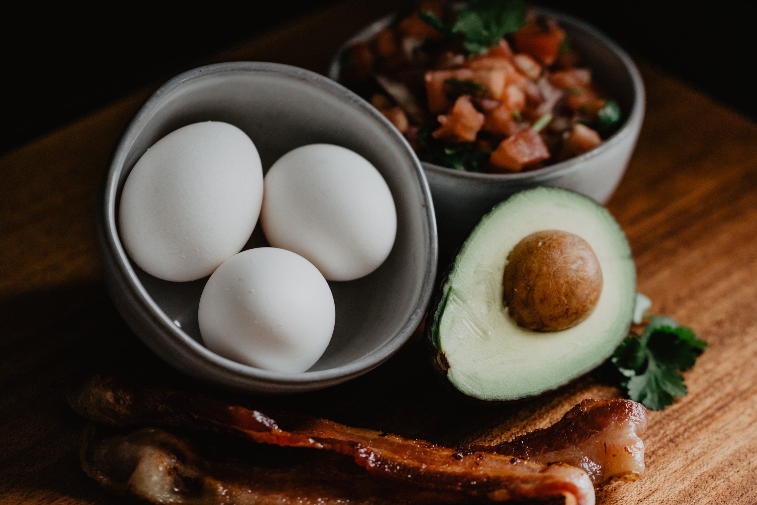 Three white eggs in a small gray bowl, half an avocado with seed showing, and a bowl of a chopped vegetable and bean salad.
