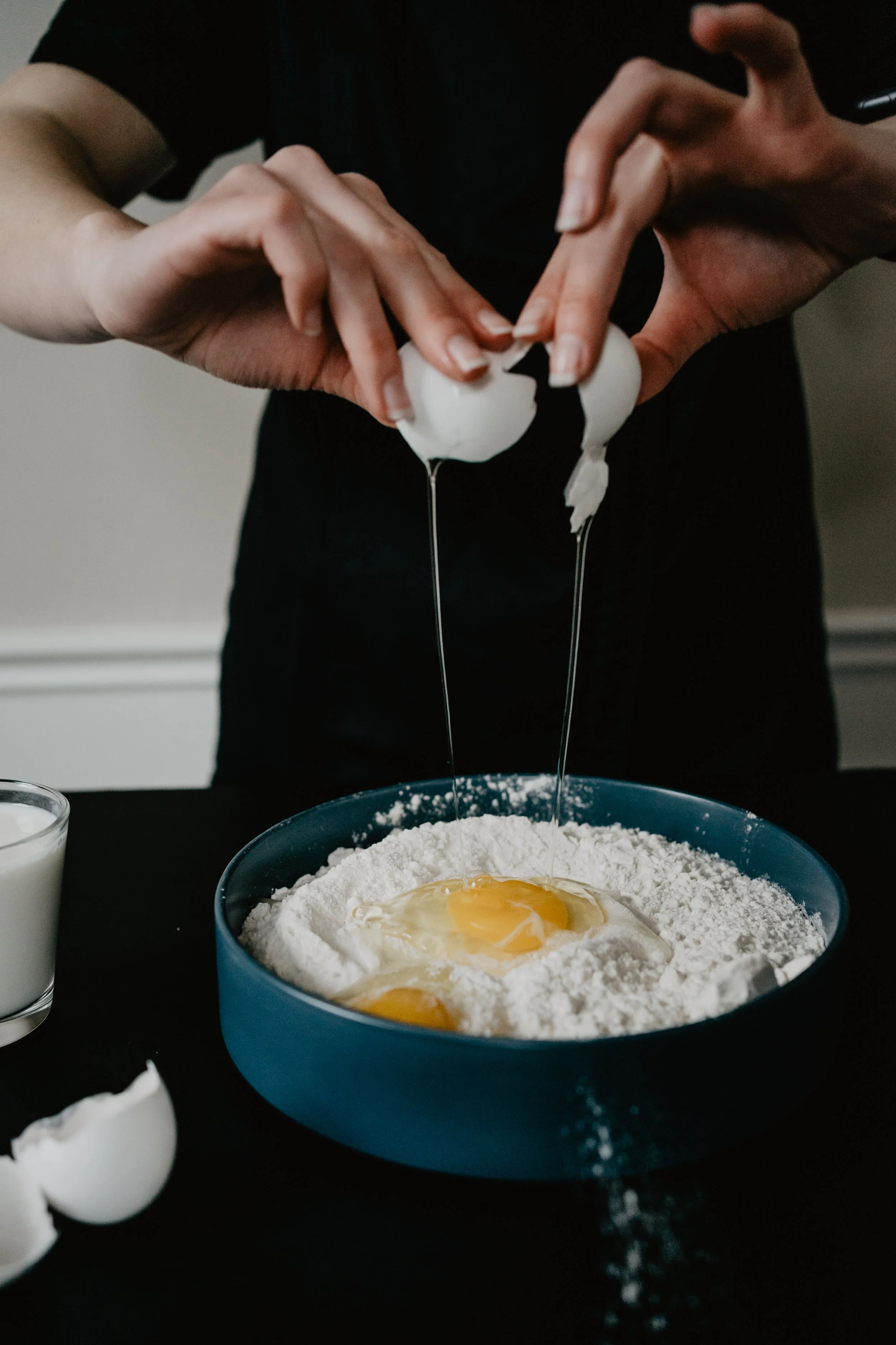 A person cracking eggs into a bowl of flour and eggs, with a glass of milk nearby on a black surface.