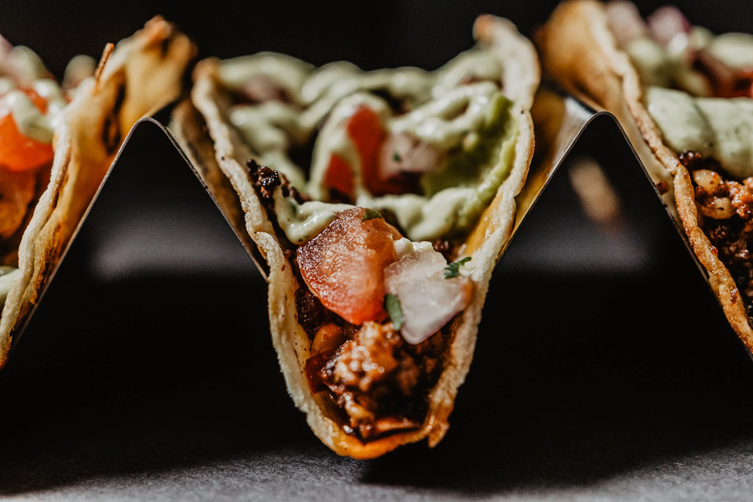Close-up of three tacos with various fillings, positioned on a dark surface.