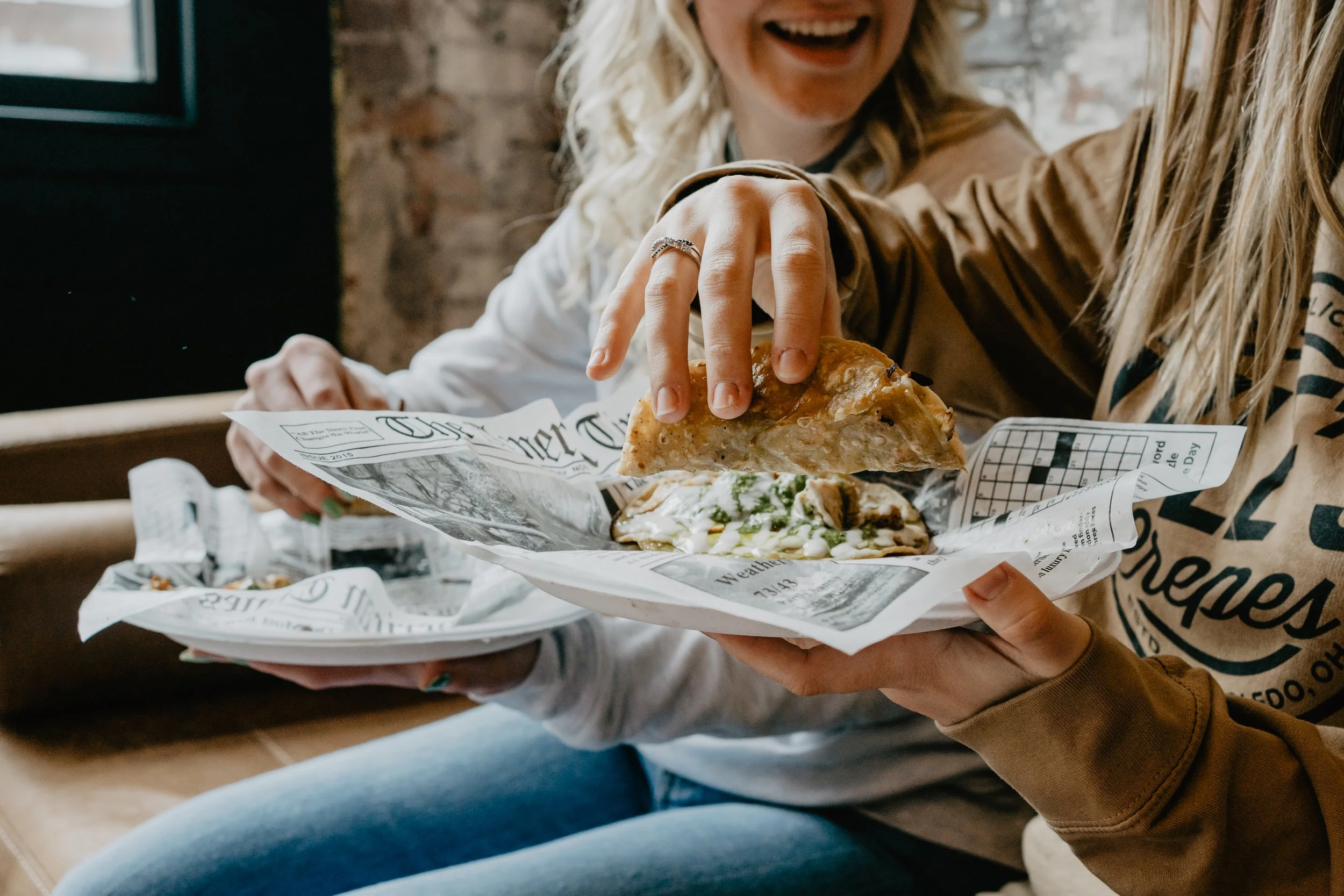 Two people holding and enjoying slices of pizza at a restaurant. The focus is on their hands and the pizza, with a brick wall background and casual attire.