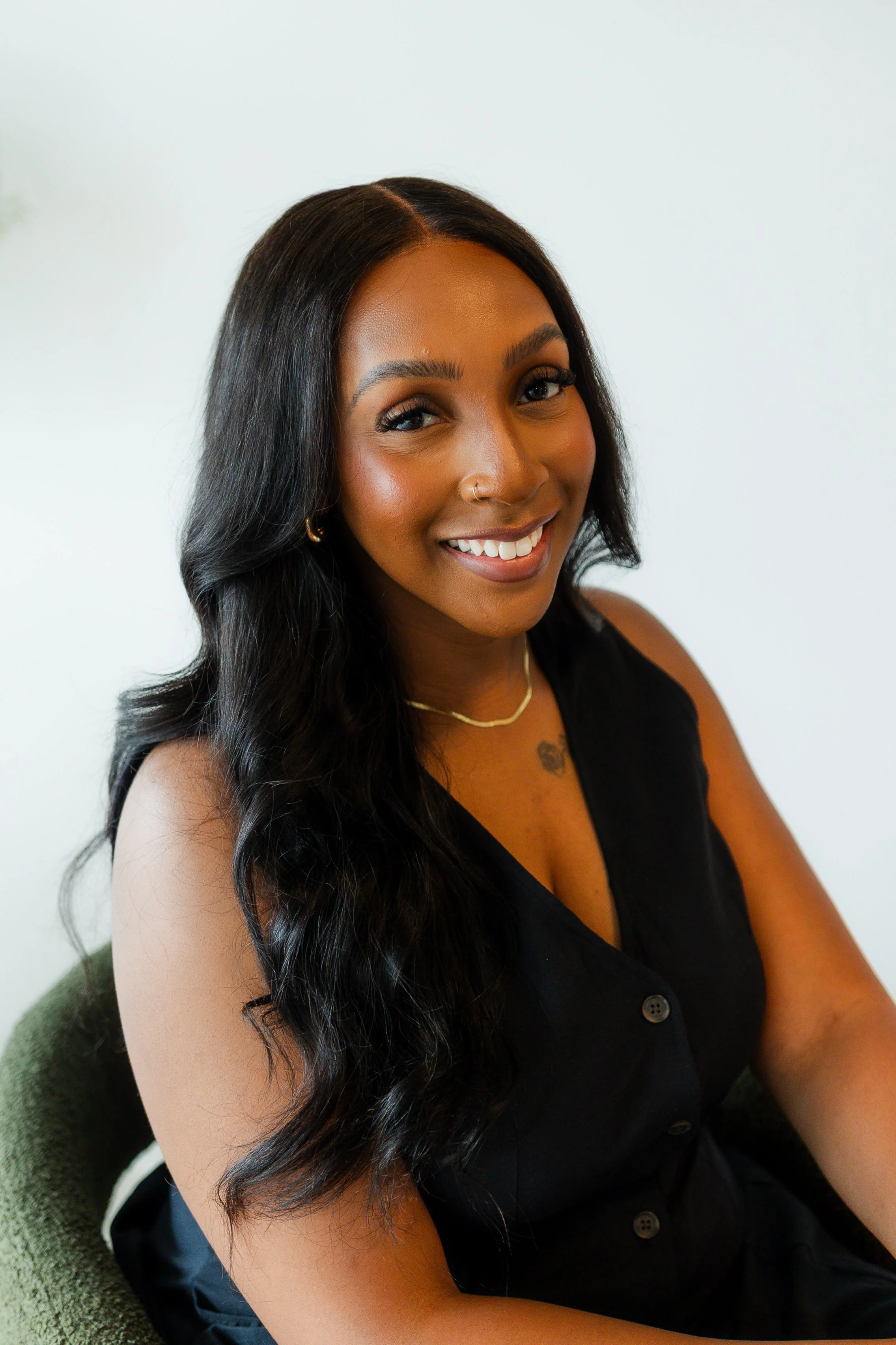 A woman with long black hair, wearing a black sleeveless top, gold jewelry, and smiling, sitting on a green chair against a white background.