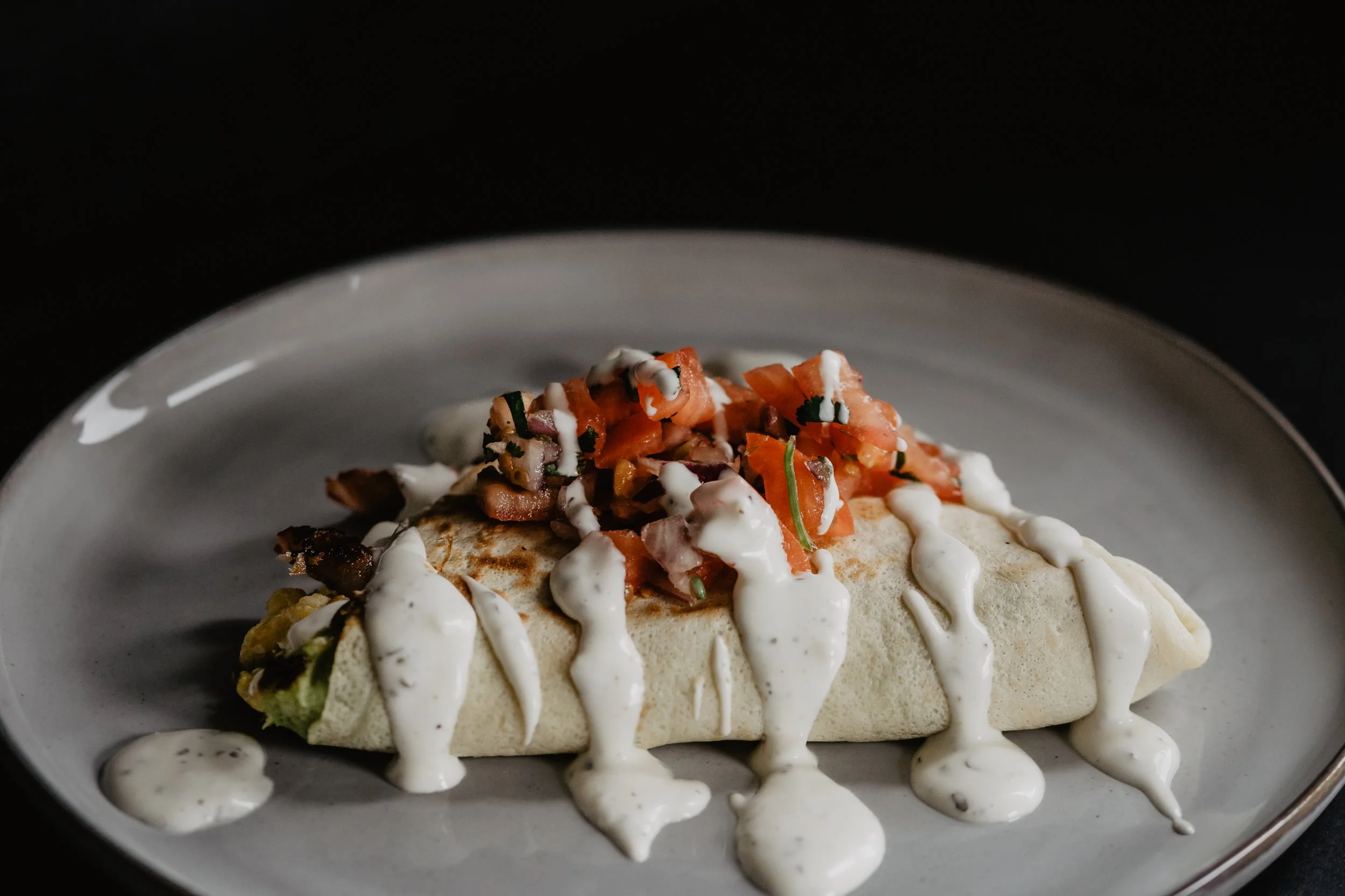 Close-up of a taco with diced tomatoes, cilantro, and white sauce, served on a white plate against a dark background.