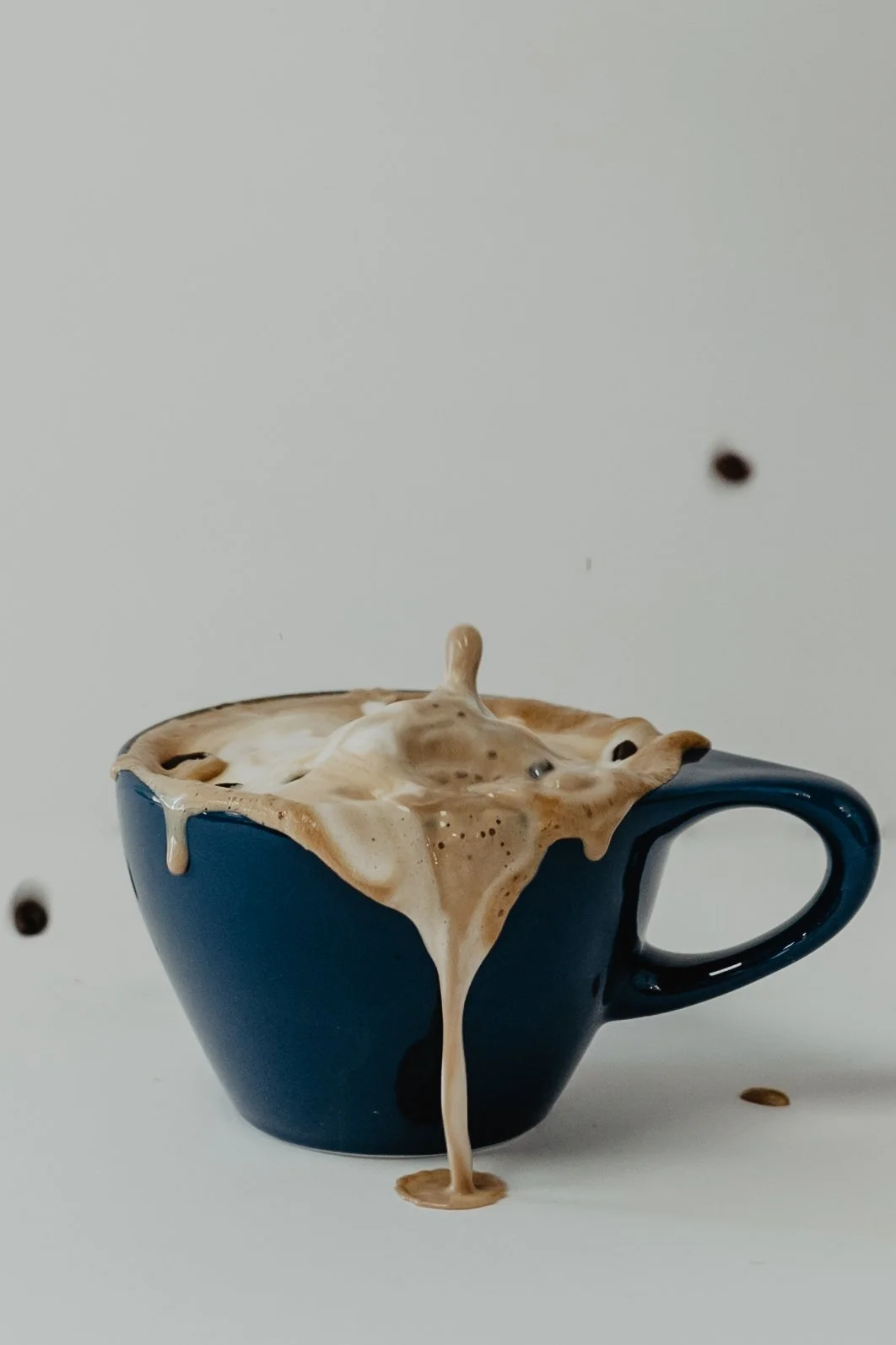 A blue mug overflowing with coffee spilling over the sides and onto the surface below, with dark and light coffee beans falling in the background.