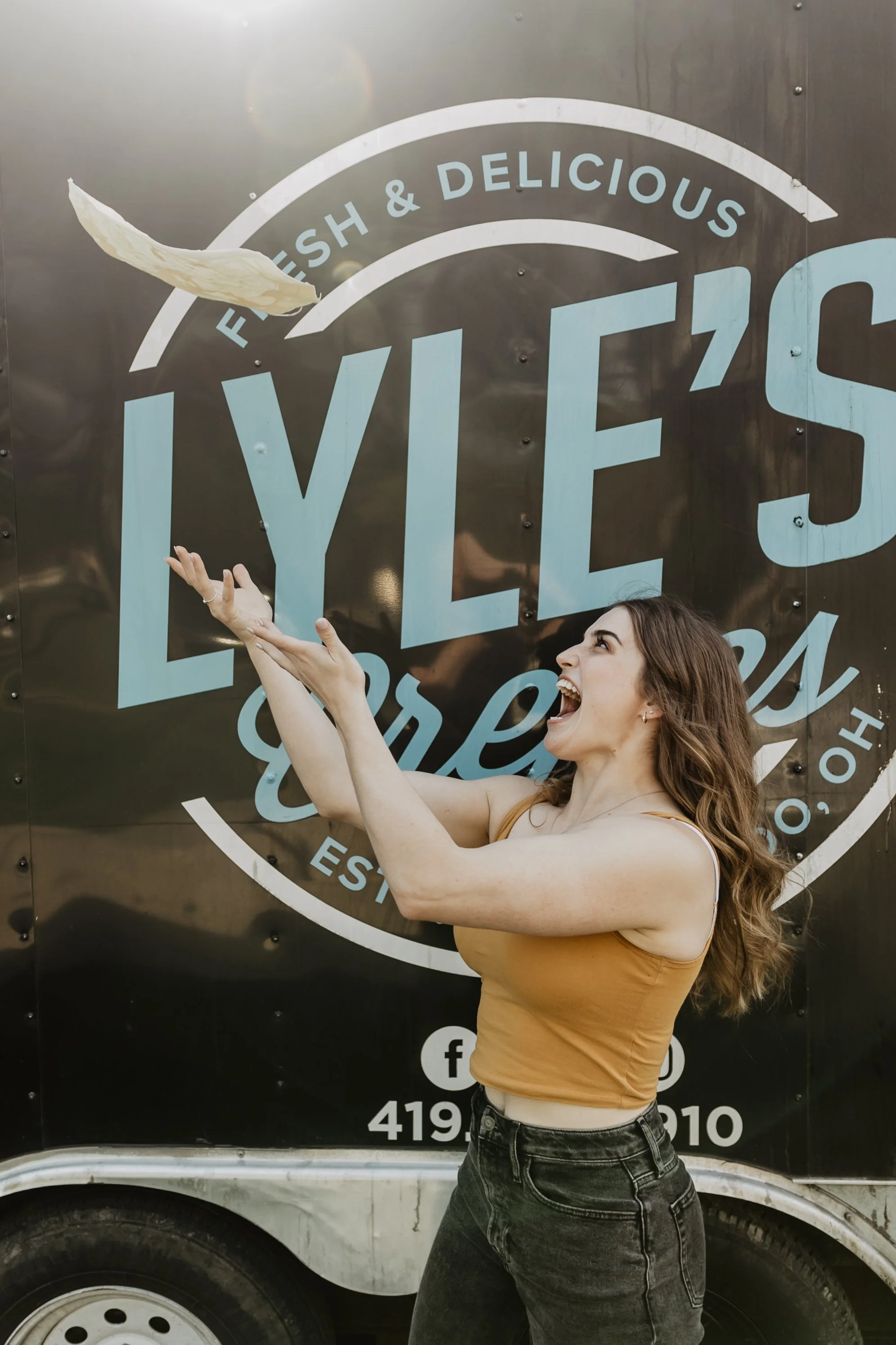 A woman with long wavy hair, wearing a tan sleeveless top and black jeans, is smiling and tossing a piece of flatbread in the air, standing in front of a black food truck with a large logo that reads 'FISH & DELICIOUS' in blue and white letters.