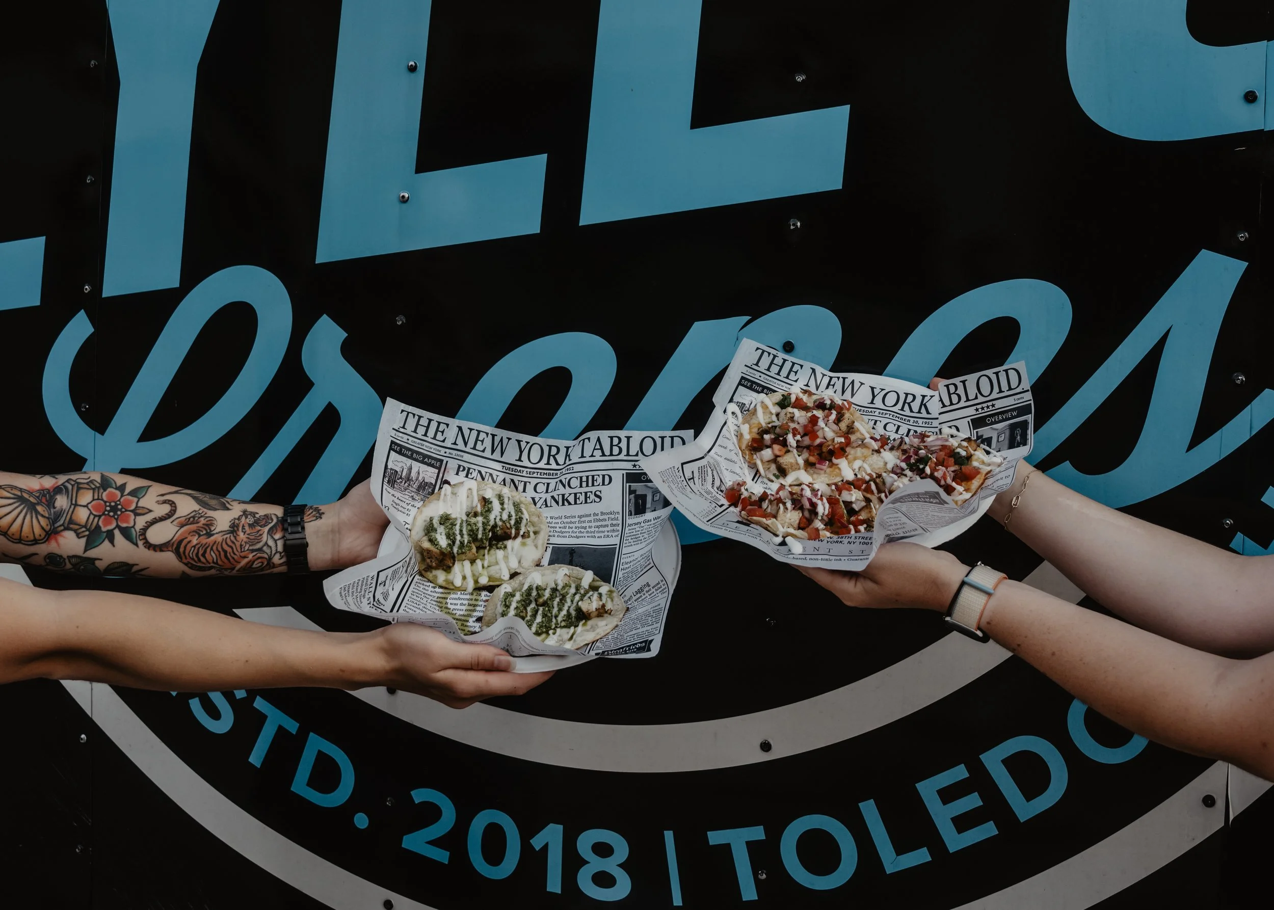 Multiple hands holding trays of food wrapped in newspaper-style paper, with one tray containing a variety of toppings and sauces, against a black background with large blue text.