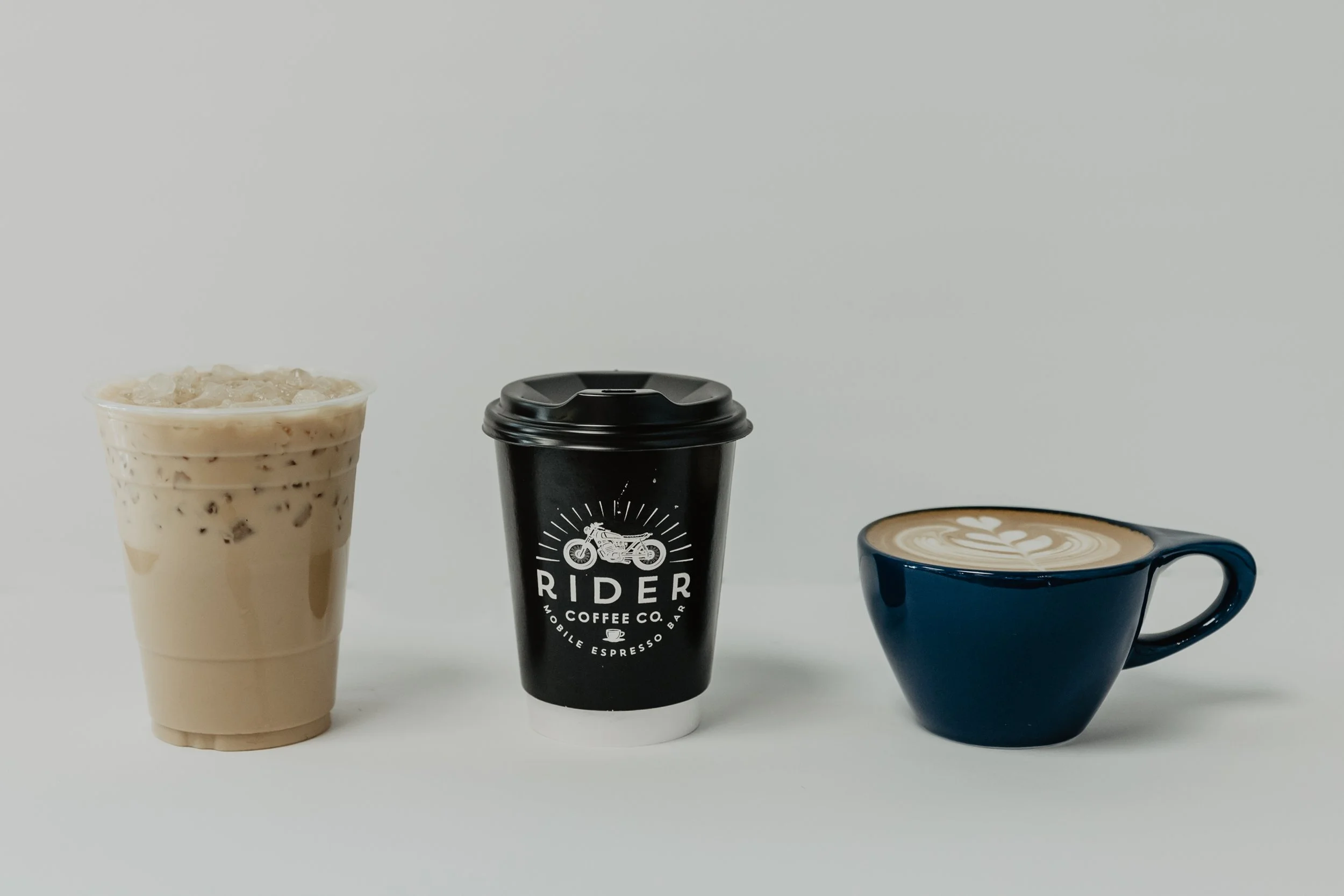 Three coffee beverages: iced coffee with ice and milk in a clear cup on the left, a black travel mug with a motorcycle logo in the center, and a latte with floral latte art in a blue ceramic cup on the right.