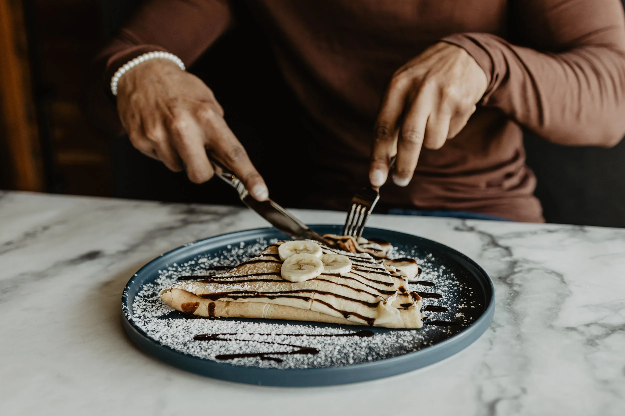A stack of crepes topped with sliced bananas, drizzled with chocolate and chocolate syrup, garnished with powdered sugar, served on a black plate on a marble table with hands using forks to cut into it.