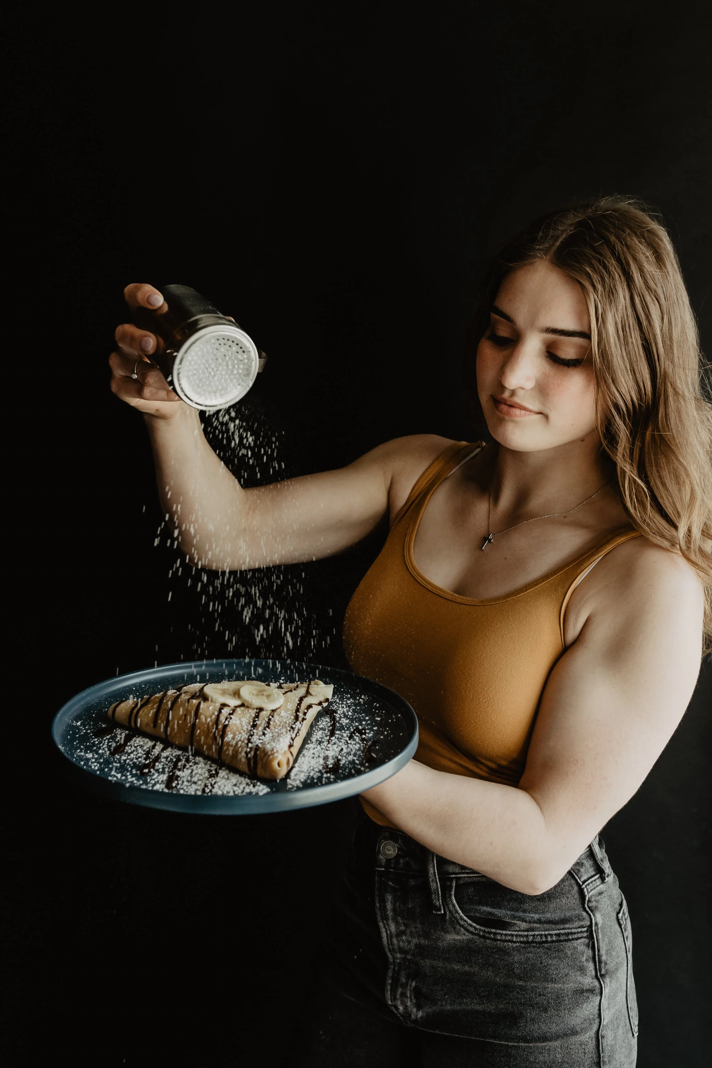 A young woman with brown hair and a tank top sprinkles powdered sugar over a dessert on a plate, with a black background.