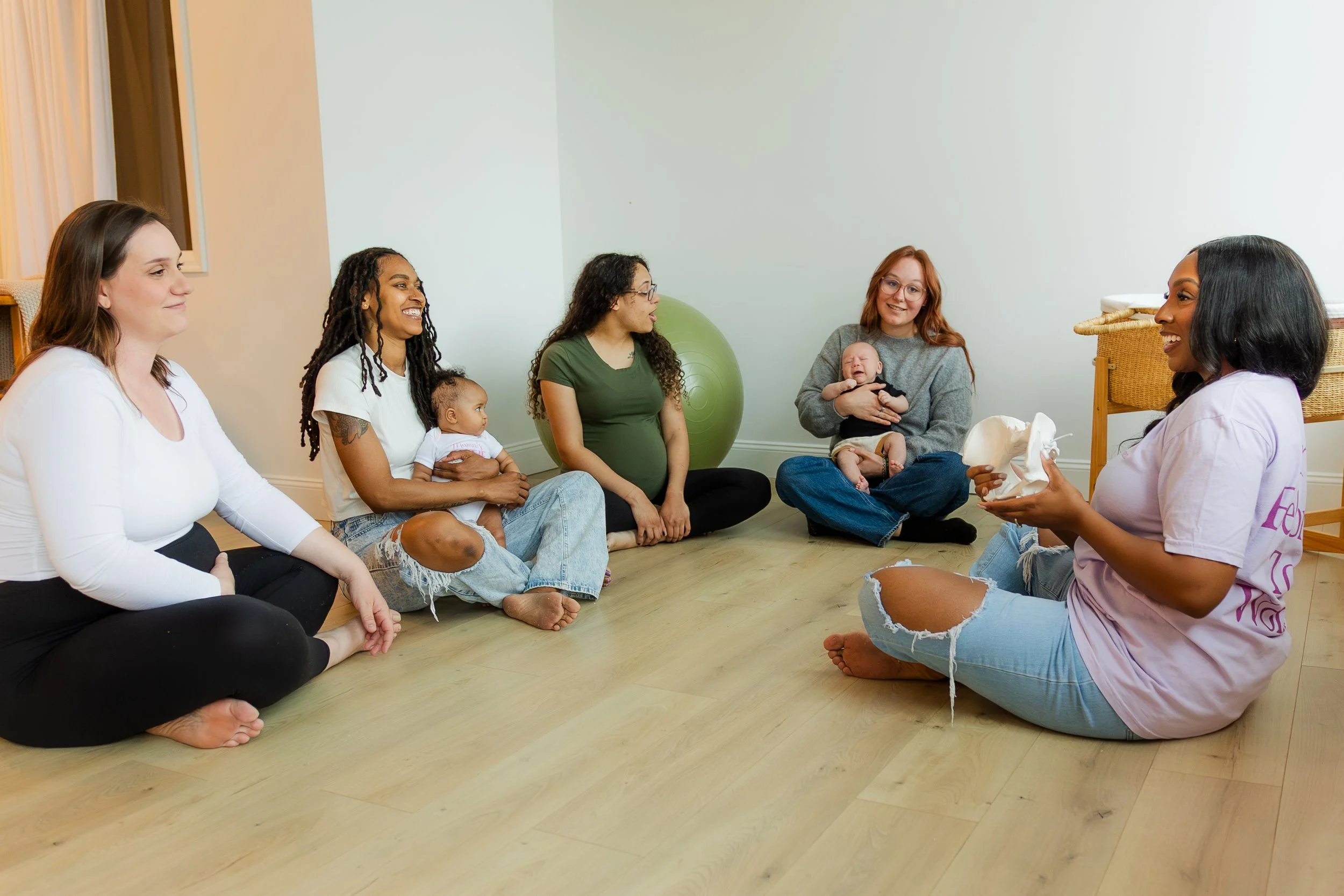 A group of women and infants sitting on the wooden floor during a discussion or class, with a pelvic model held by one woman.