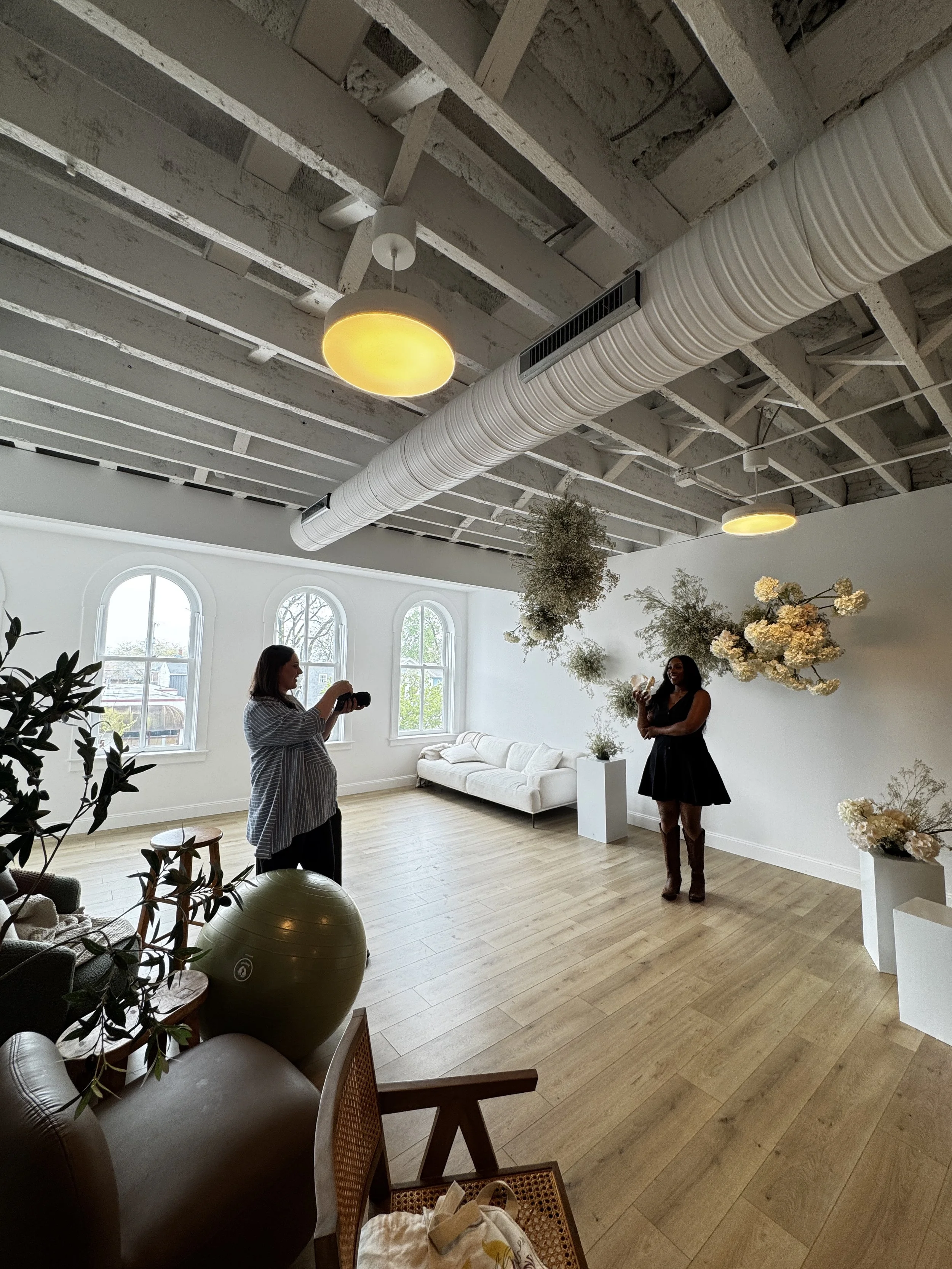Two women are in a bright, modern room with large arched windows, light wood floors, and decorative white flowers on the wall. One woman is wearing a black dress and brown boots, smiling and holding flowers, while the other woman is taking her photo. The room has exposed ceiling beams and a large white ventilation duct.