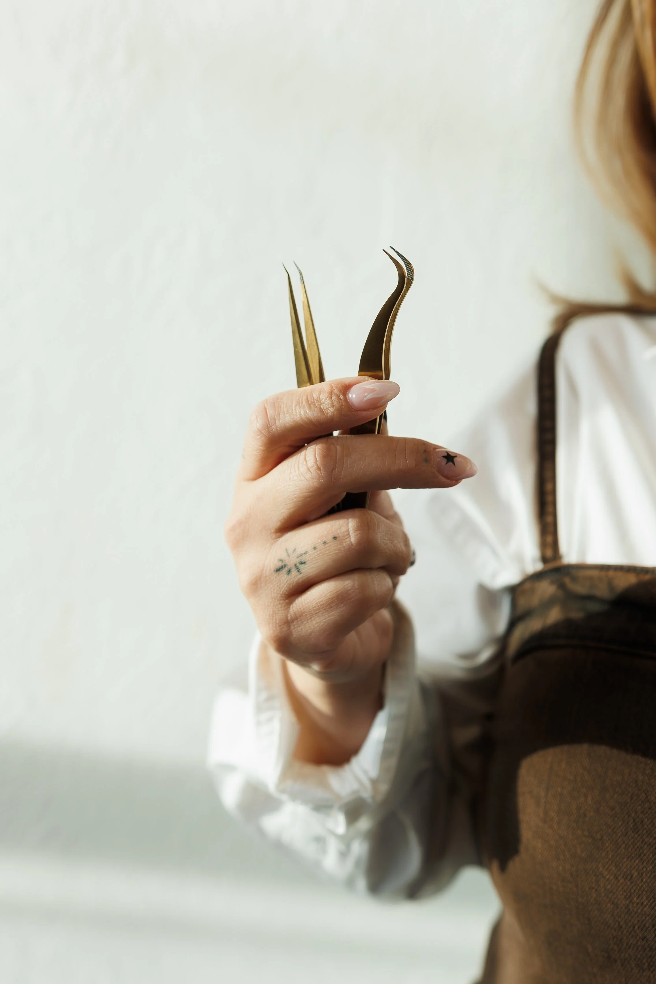 Close-up of a woman's hand holding two small tweezers, with a tattoo on her middle finger and a star tattoo on her ring finger, against a plain white background. She is wearing a white shirt and brown vest.