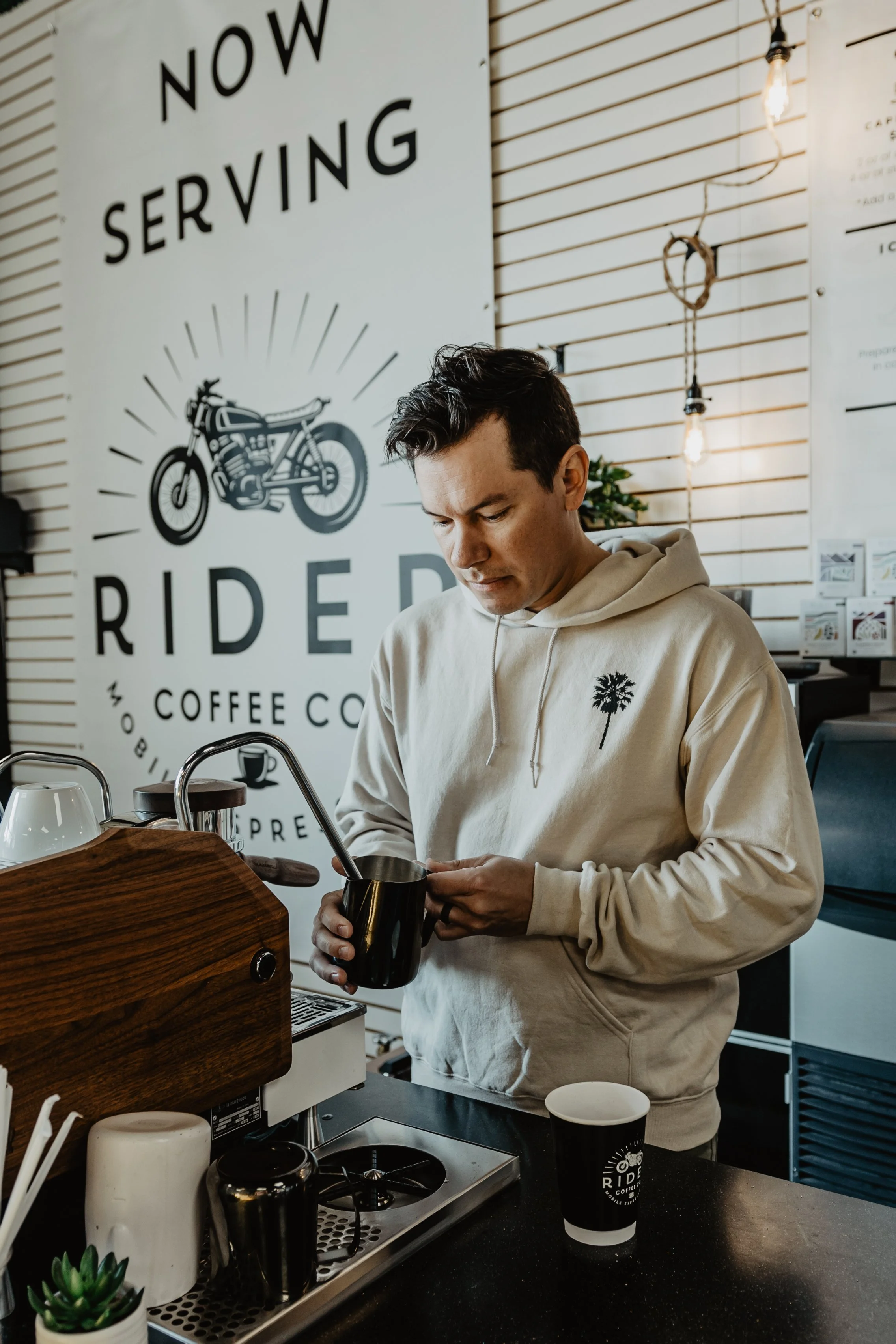 A man with dark hair wearing a beige hoodie with a palm tree logo is preparing a drink at a coffee shop counter. Behind him is a large sign with the words 'Now Serving' and an illustration of a motorcycle, along with the words 'Ride' and 'Coffee'.