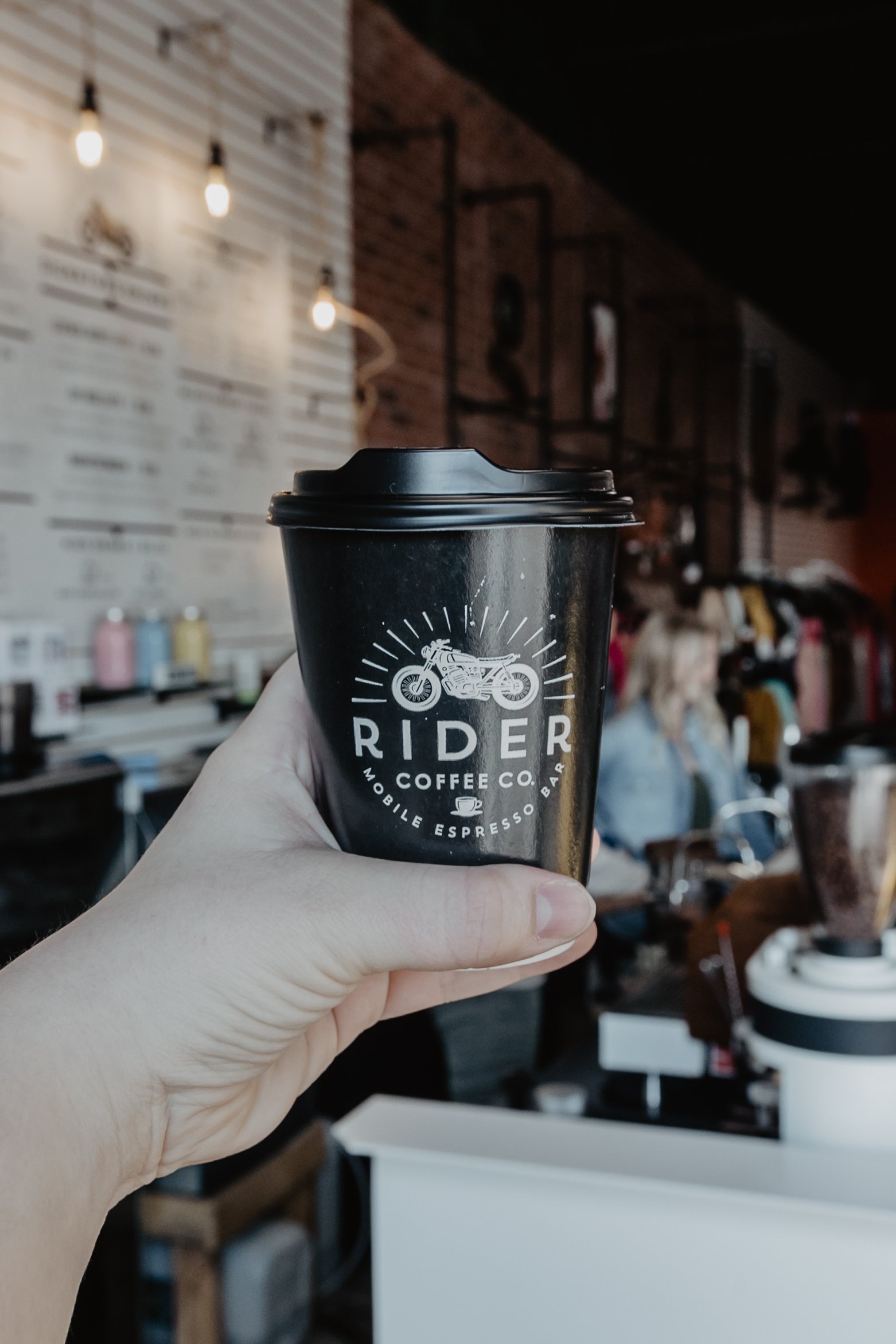 Hand holding a black coffee cup with a motorcycle logo and the words 'Rider Coffee Co.' inside a cozy cafe with warm lighting and decorative shelves.
