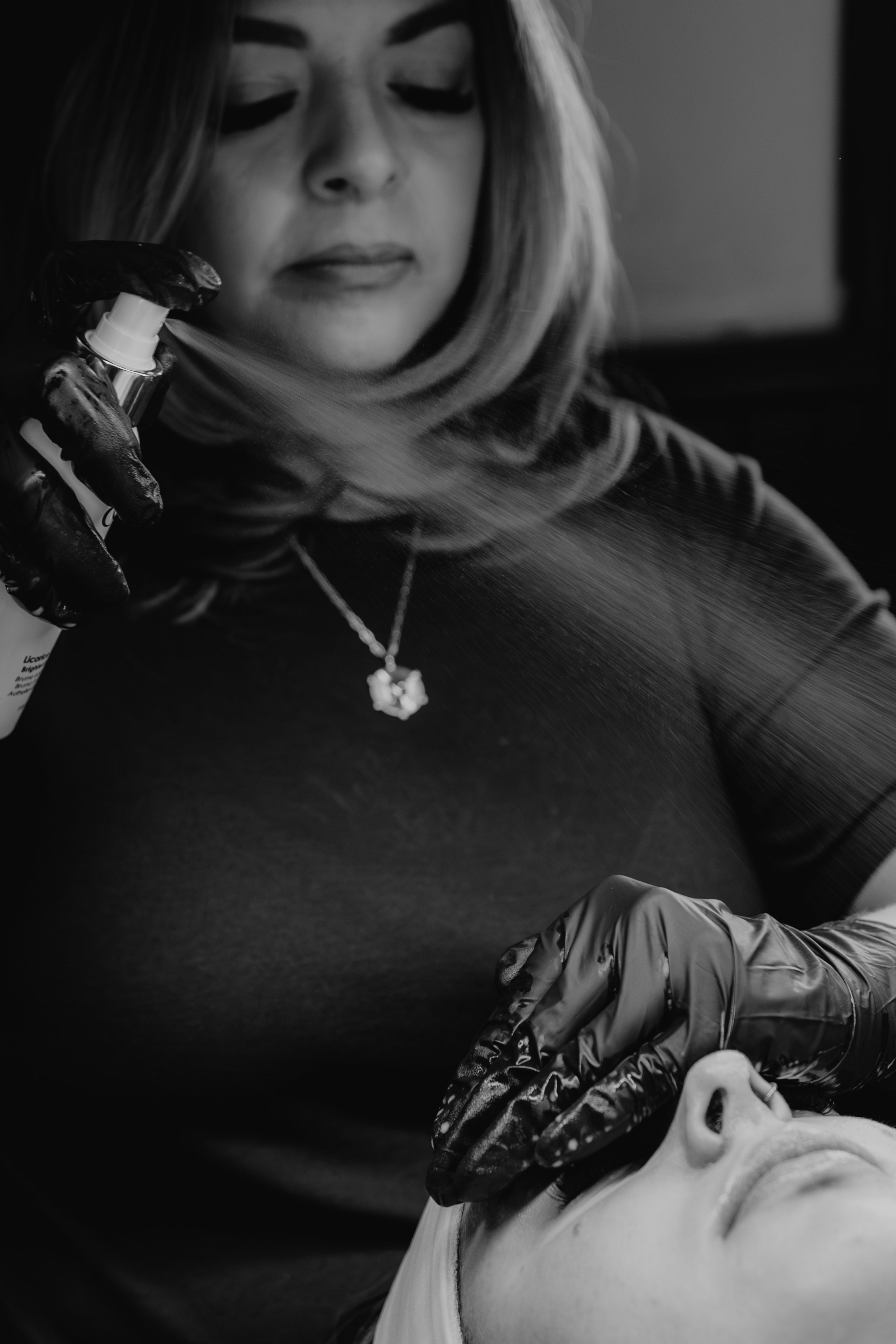 A woman lying on her back with her eyes closed, receiving a spray or mist from a person wearing black gloves, in a monochrome setting.